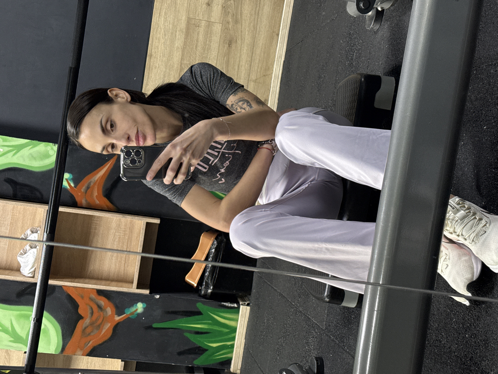 An indoor mirror selfie captures a female in a fitness facility located in Pleven, Bulgaria. The subject is a young adult female with dark hair pulled into a ponytail, wearing a grey t-shirt, light purple/white wide-leg trousers, and white athletic shoes. She is seated on a black, multi-tiered fitness step bench, holding a dark-colored smartphone in her right hand to take the photograph. Her left forearm displays a tattoo, and her left wrist has multiple bracelets. The flooring is black rubber gym matting. The background wall features a dark base with a vibrant mural depicting stylized green foliage and orange organic shapes. Integrated into the wall are light wooden cubbies, one containing a crumpled white item of clothing. A black trash can with a brown lid is visible on the floor near the wall. A horizontal grey bar and a vertical silver cable, likely components of gym equipment, are visible in the foreground's reflection.