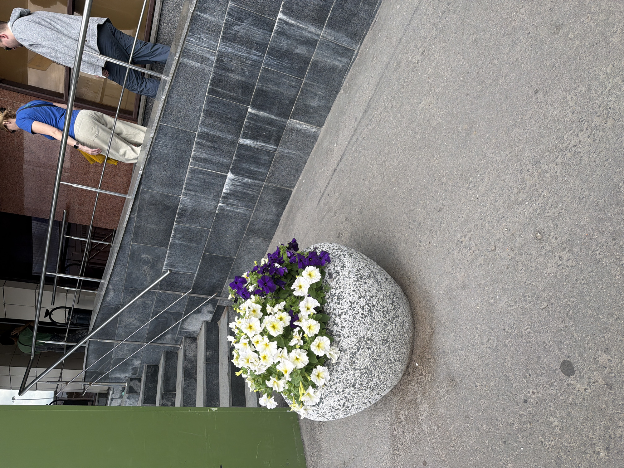 A flower pot filled with white and purple petunias sits on a concrete sidewalk in front of a building. The building has a stone facade and a set of stairs leading up to an entrance. Two people are walking up the stairs. One person is wearing a blue shirt and light-colored pants, and the other is wearing a gray hooded sweatshirt and dark pants. The weather appears to be overcast.