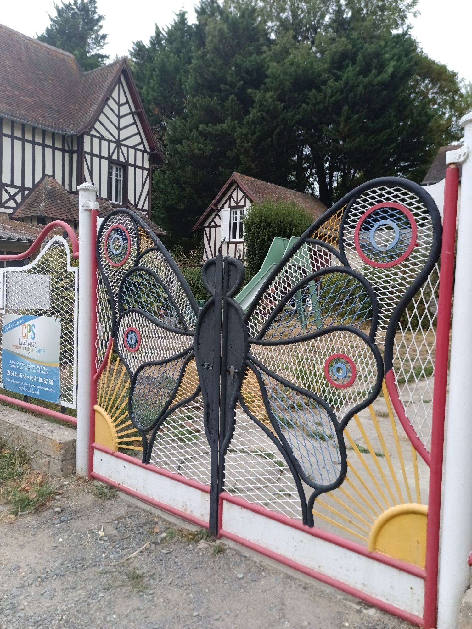The image shows a decorative metal gate in the shape of a butterfly, located in front of a building. The building appears to be a house with a distinctive architectural style, featuring white walls with dark timber framing and a brown tiled roof. Behind the house, there is a dense stand of trees. The gate itself is elaborate, with intricate metalwork forming the butterfly's wings, which are decorated with circular patterns in red, blue, and yellow. There is also a playground visible behind the gate, including a green slide. A sign is attached to the left side of the gate, providing information about a company called "CPS Energies". The ground in front of the gate is covered with gravel and dirt, with some dry leaves and vegetation. The overall scene suggests a residential or recreational area in a suburban or rural setting. The building's architecture indicates it might be in Normandy, France, and the sign is in French, suggesting the location is Houlgate, France.