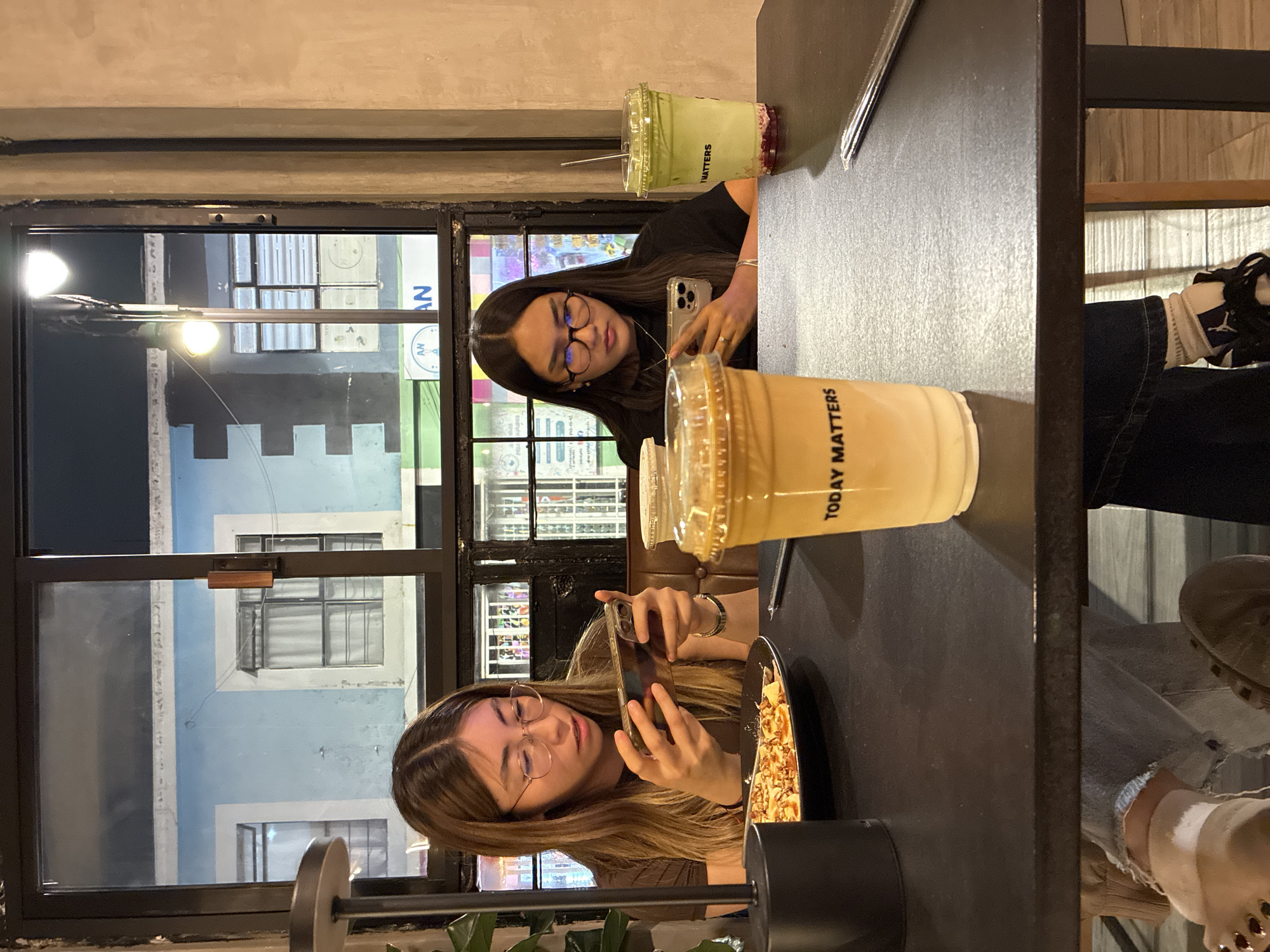 Two young women are seated at a dark table in a restaurant. Both are using their smartphones. The woman on the left is photographing a plate of food. The woman on the right is looking at her phone. On the table are two drinks, one green and one beige, in clear plastic cups with the text "TODAY MATTERS" printed on them. Behind them, through a large window, the exterior of a building is visible. The building is light blue with white trim and appears to be located in San Andrés Cholula, Mexico, based on architectural characteristics.