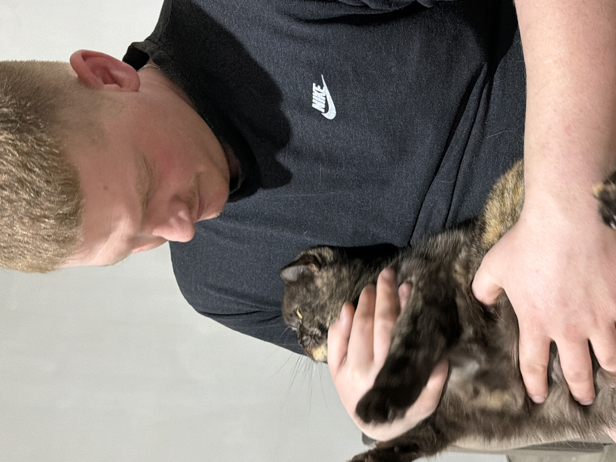 The image shows a person with short blonde hair wearing a black long-sleeved Nike shirt, holding a tortoiseshell cat. The person is looking down at the cat, who appears to be held securely in the person's hands. The background is a plain, light gray or white wall. There is no indication of the location being Chaikovsky, Russia, or any specific city.