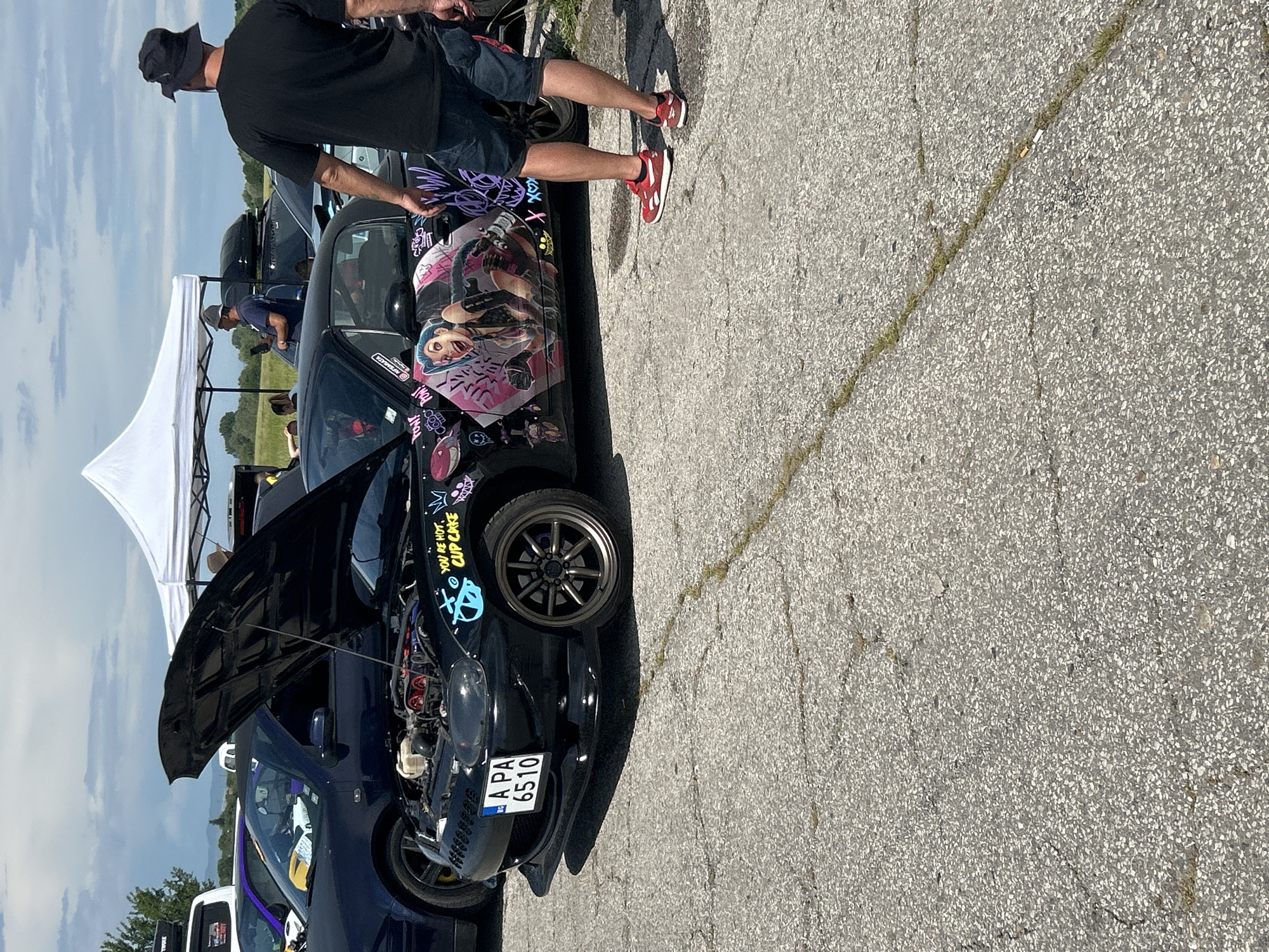 The image shows a black car with its hood open, displaying engine components, at an outdoor event, likely a car meet, in Dalbok Izvor, Bulgaria. The car features custom paint and decals, including a cartoon character on the door. A man in a black shirt and bucket hat stands next to the car on the passenger side. Part of a white tent is visible in the background. The car has a license plate "A PA 6510," indicating registration in Bulgaria. The ground is paved with cracked concrete.