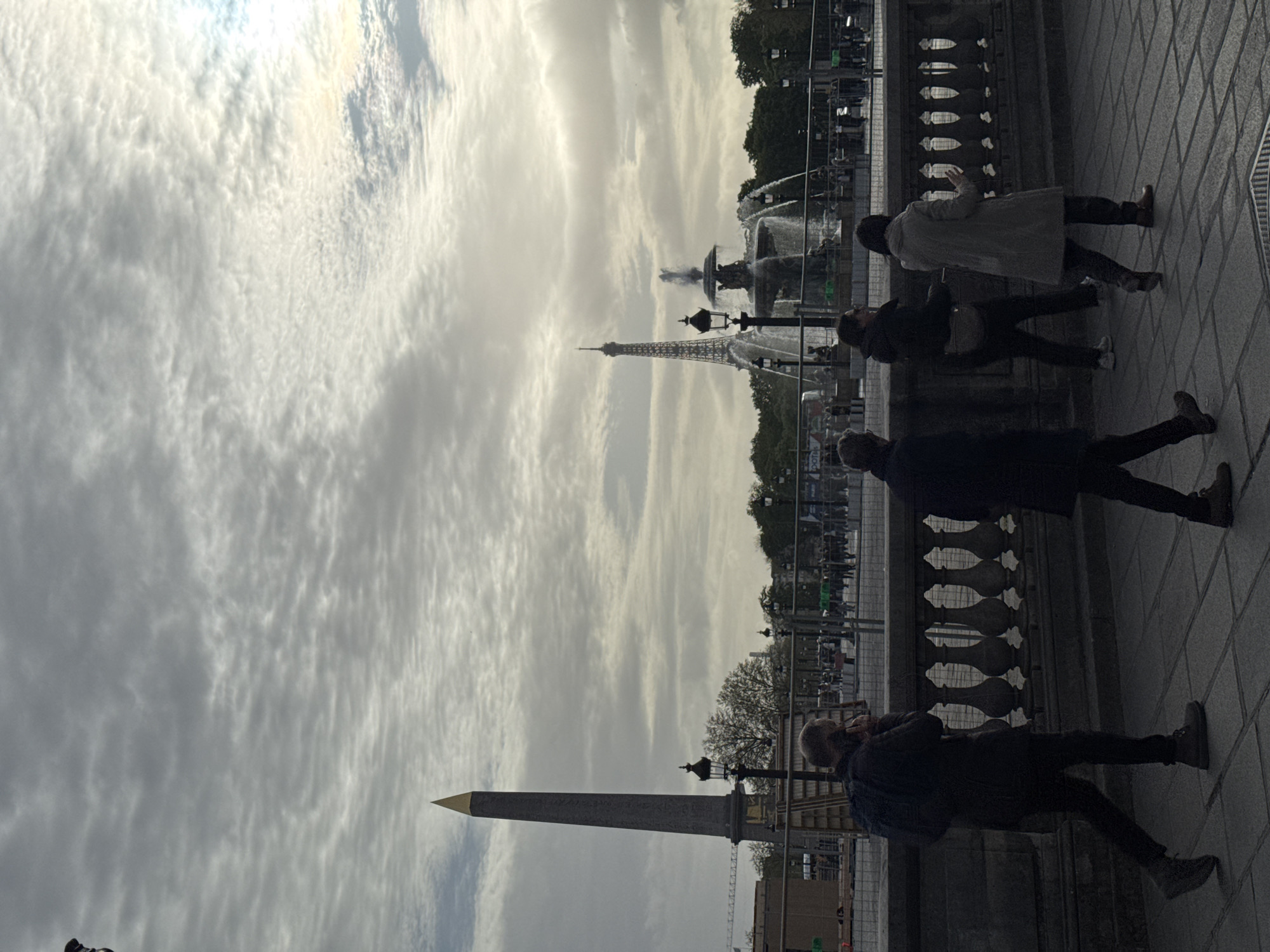 The photograph, taken in Paris, France, shows a view of the Place de la Concorde and the Eiffel Tower in the background. In the foreground, the Luxor Obelisk stands prominently. A fountain is situated mid-ground, surrounded by a fence and people. Several pedestrians are walking along a stone walkway. The sky is overcast with a layer of textured clouds. The image is oriented sideways.