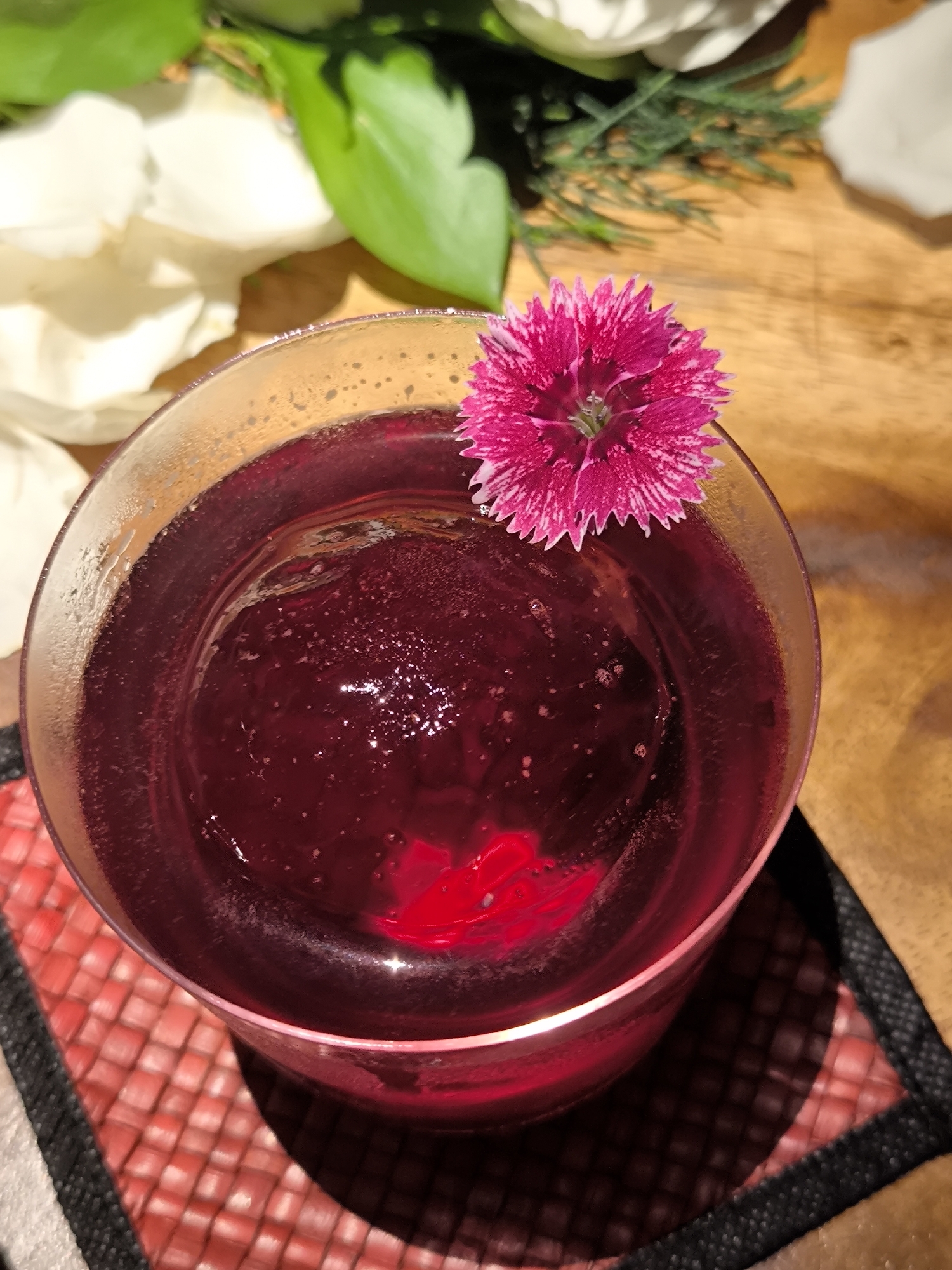 The image displays a close-up, top-down view of a vibrant, deep reddish-purple drink in a clear glass, set in North Kuta, Indonesia. A large, spherical ice cube is submerged in the liquid, which shows small bubbles on its surface. The rim of the glass exhibits subtle condensation.
A striking fuchsia-pink flower, possibly a dianthus or miniature carnation, with delicate white speckled patterns and fringed petals, is perched as a garnish on the glass's rim. The glass rests on a square coaster, which features a woven, basket-weave pattern in red hues, framed by a black border.
The background elements contribute to a natural and inviting scene. To the left, soft white petals and lush green leaves are visible, while to the right, further green foliage, resembling pine needles, and a warm wooden surface complete the setting. The lighting is soft and ambient, highlighting the textures of the flower and the condensation on the glass. The scene evokes a sense of relaxation and enjoyment, likely featuring a refreshing beverage such as a cocktail. No people or visible text are present.