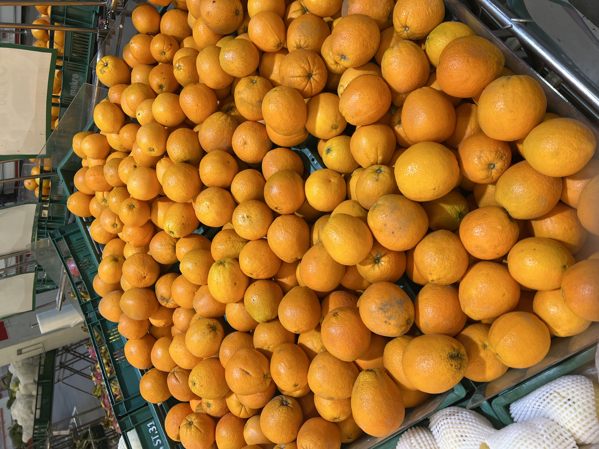 An overhead shot showcases a vibrant display of fresh oranges neatly arranged in green plastic crates within a supermarket. The crates are filled to the brim with a multitude of round, citrus fruits, their bright orange hues creating a striking visual contrast against the dark green plastic. Some of the oranges show slight imperfections, such as minor blemishes or variations in color, which are typical of fresh produce. The background is blurred, but hints at more produce and shelving, suggesting a bustling market environment. A label with "ST.31" is visible on one of the crates, indicating a product code or section. The overall scene is clean and organized, highlighting the abundance and freshness of the oranges available for sale at the supermarket. This location is likely in Baan Bo Phut, Thailand.
