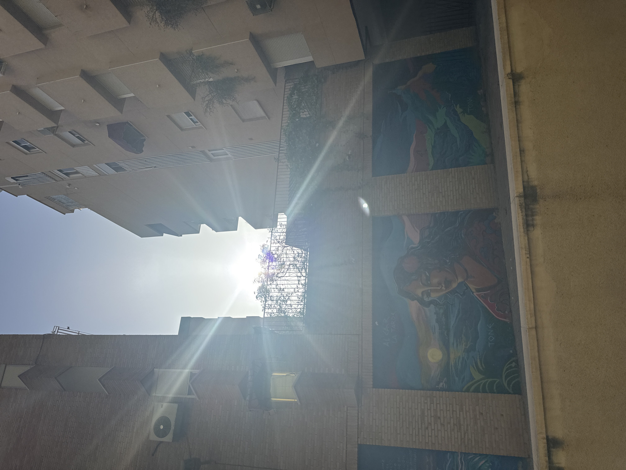 The image shows a narrow street between two apartment buildings, with the sun shining brightly overhead. A mural is painted on the lower part of the left building, depicting a woman with long, dark hair and a serene expression, set against a backdrop of water and a sunset. The text "ALCAR LA TARDE" and "TON" are visible on the mural. The buildings are made of brick and concrete, with balconies and windows. An air conditioning unit is visible on the left building. The street appears to be located in the city of Murcia, Spain, given the context provided by the mural's text.