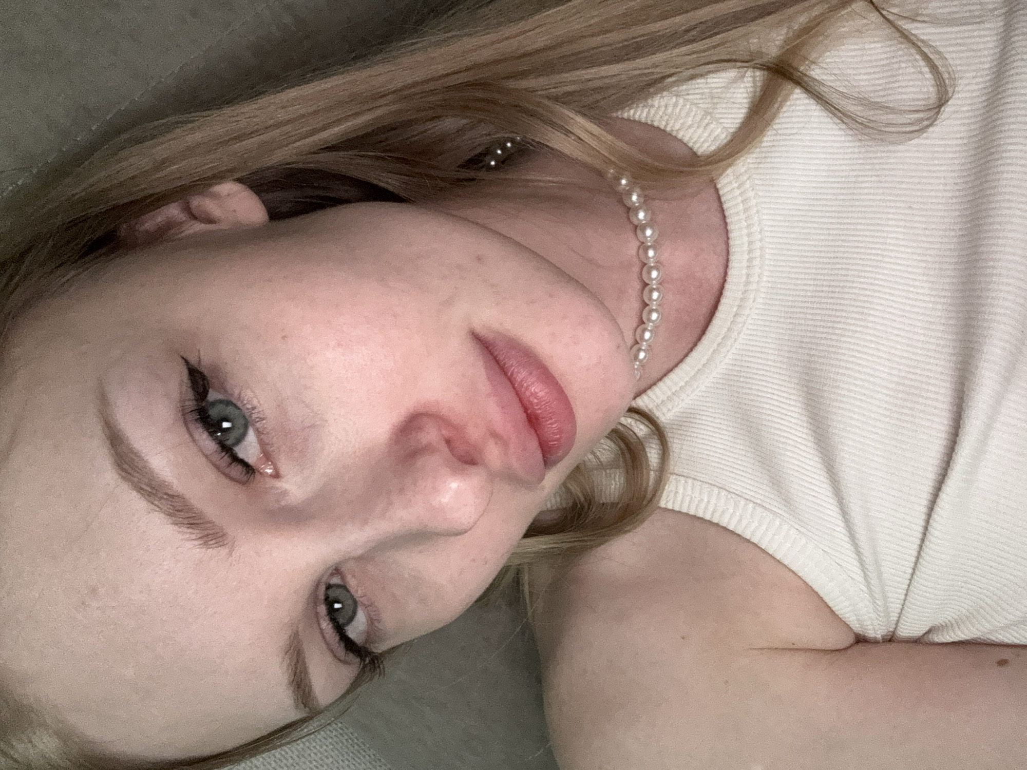 The image shows a close-up selfie of a young woman with blonde hair, blue eyes, and light skin. She is wearing a white tank top and a pearl necklace. Her makeup includes eyeliner and mascara. The background appears to be an indoor setting, possibly a couch or bed, with a textured surface visible. The image is a portrait style with the subject looking directly at the camera. There is no discernible information in the image that allows for the identification of its location within the city of Perm, Russia.