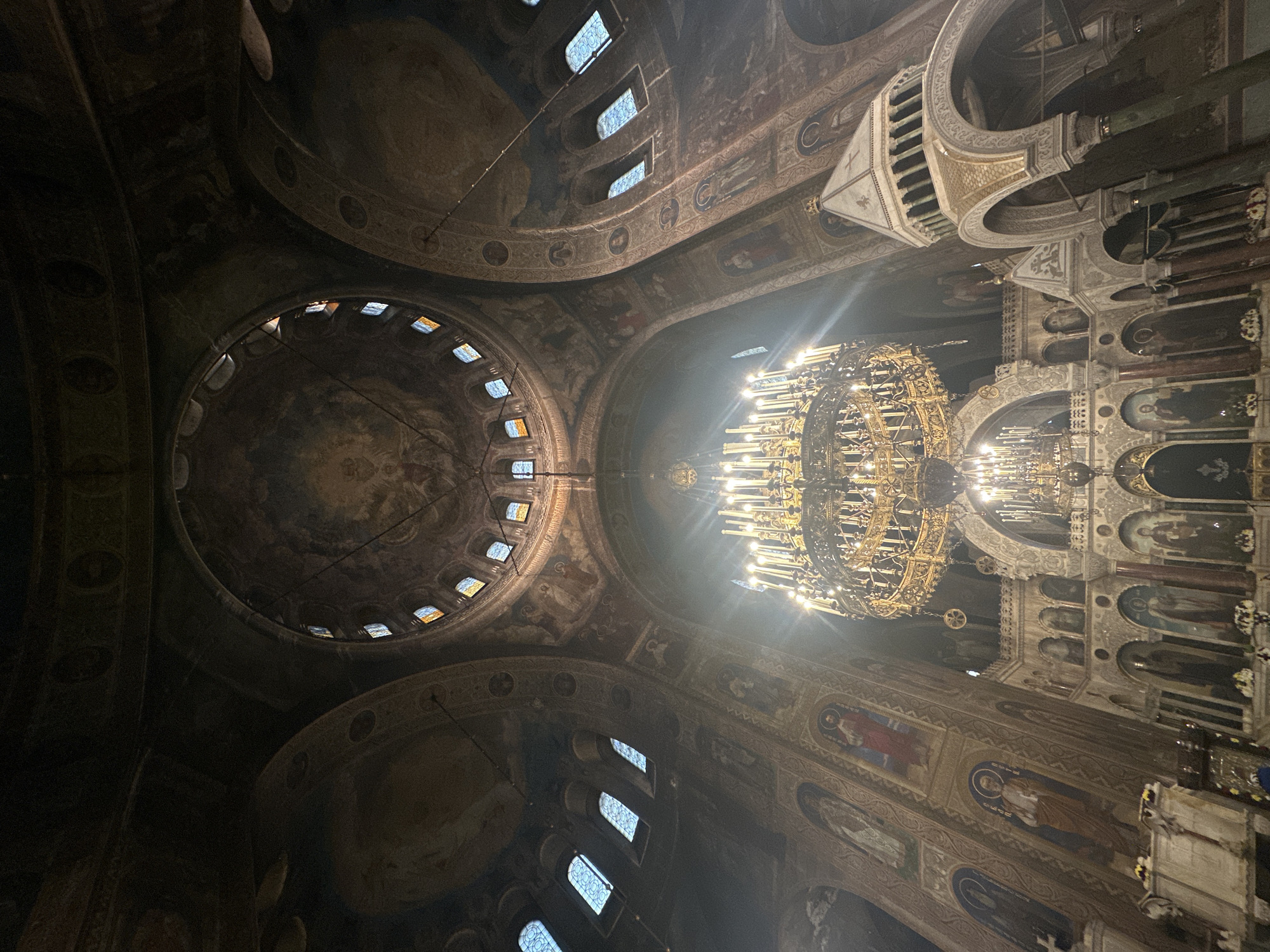 The image depicts the interior of a church in Sofia, Bulgaria. The viewpoint is angled upwards towards the ceiling, showcasing the architecture and ornamentation. A large, ornate chandelier is suspended from the ceiling, illuminated and appearing quite bright in contrast to the darker surroundings. The ceiling is adorned with frescoes and features a central dome with small windows. Arches and columns are visible, decorated with religious imagery. The overall environment suggests a historic, religious setting.