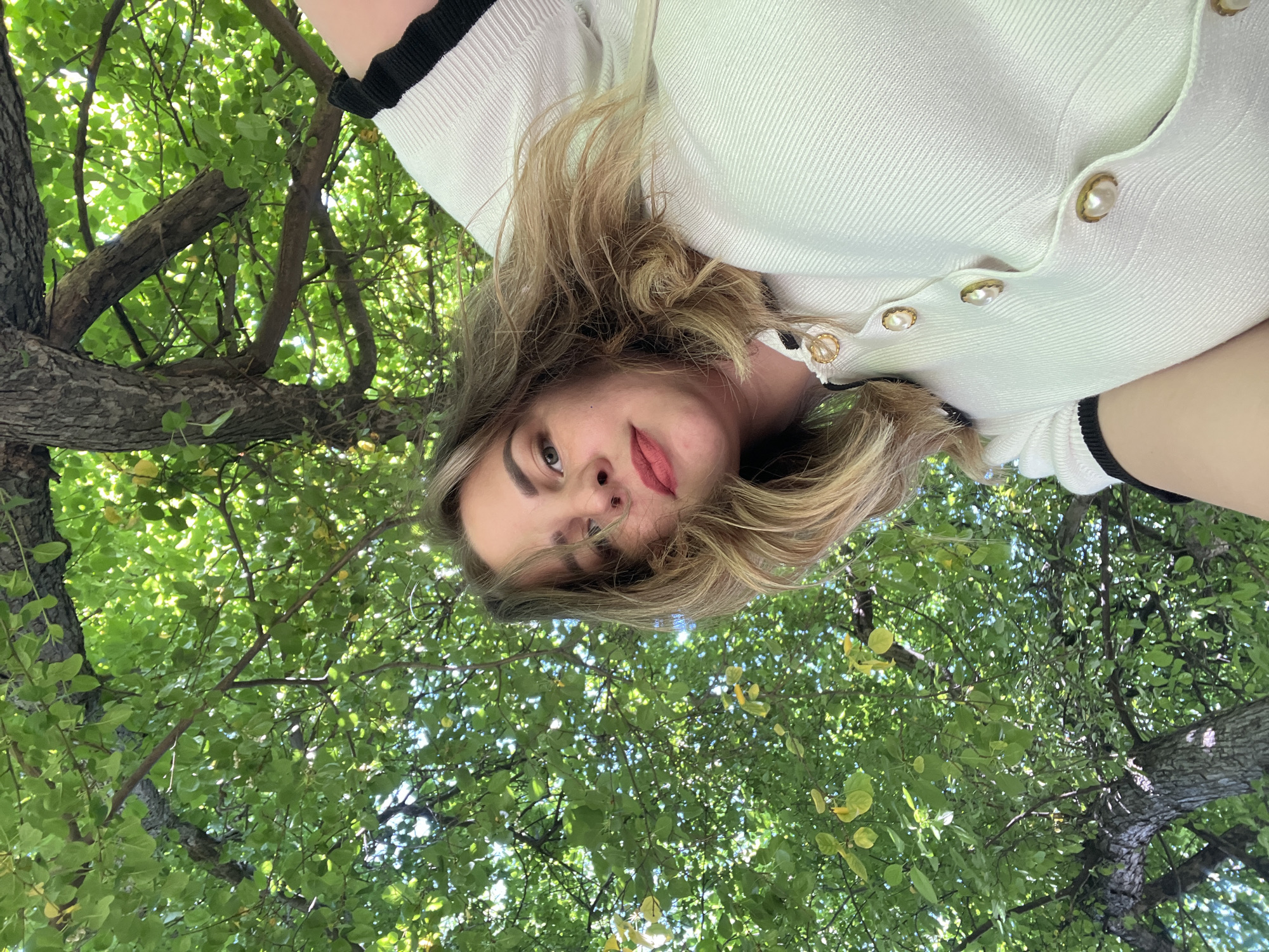 A person with fair skin, light brown hair, and wearing a white top with black trim is looking down at the camera from within the branches of a tree with green leaves. The leaves create a dense canopy above and around the person. The exact location in the city of Izhevsk, Russia cannot be determined from the image.