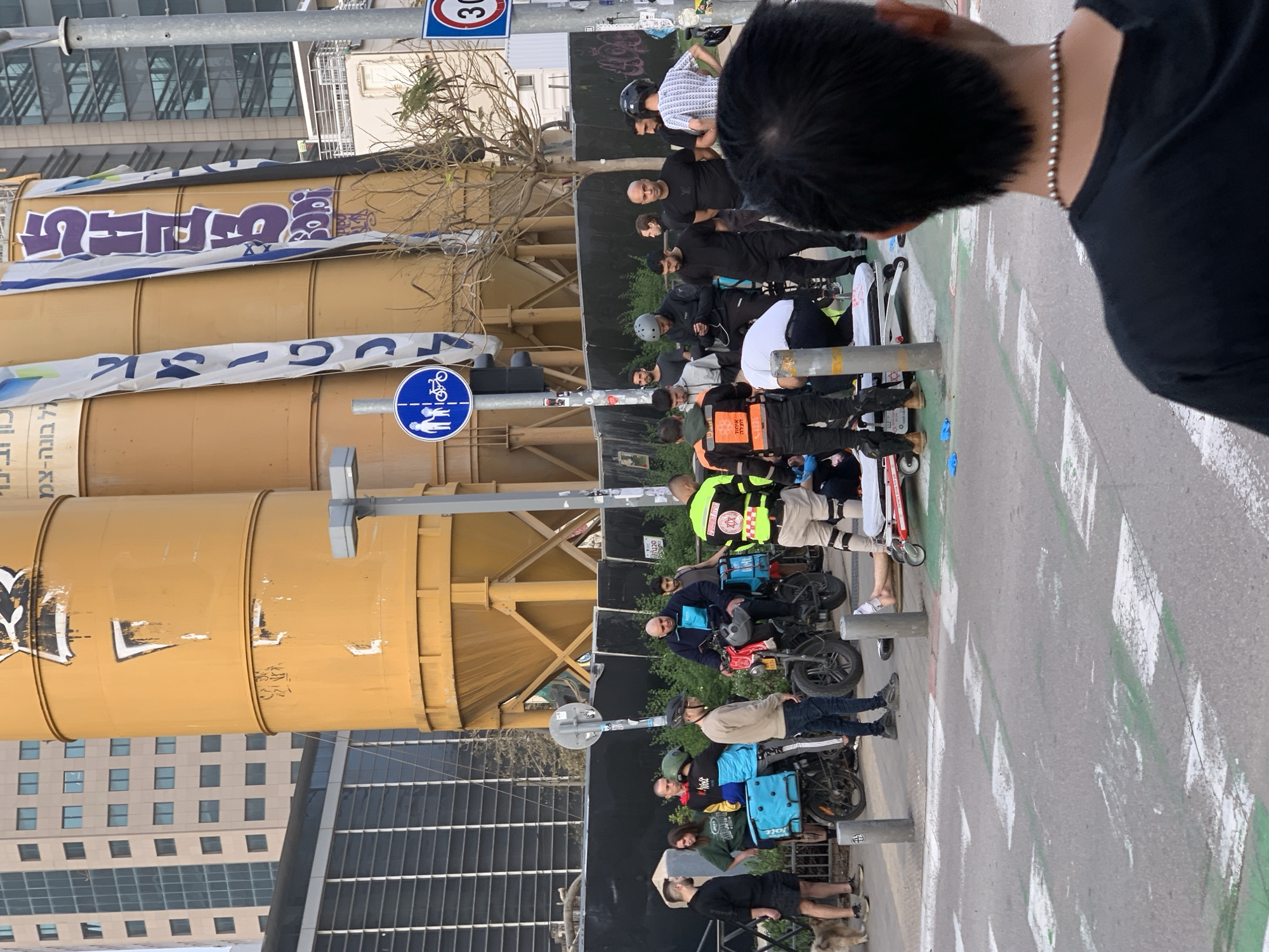 The image shows a group of people standing on a city street, likely in Tel-Aviv, Israel, given the building architecture and signage visible. The people are of diverse ages and appearances, some wearing helmets and delivery gear, suggesting they may be delivery workers. They are positioned near a large, cylindrical structure that appears to be part of a construction site or industrial facility. Street markings, including pedestrian crossing lines and a bicycle lane symbol, are visible on the asphalt road. A traffic sign with a "30" speed limit is seen in the background. Buildings with a mix of architectural styles are visible in the distance, including modern glass structures and older, residential-style buildings.
