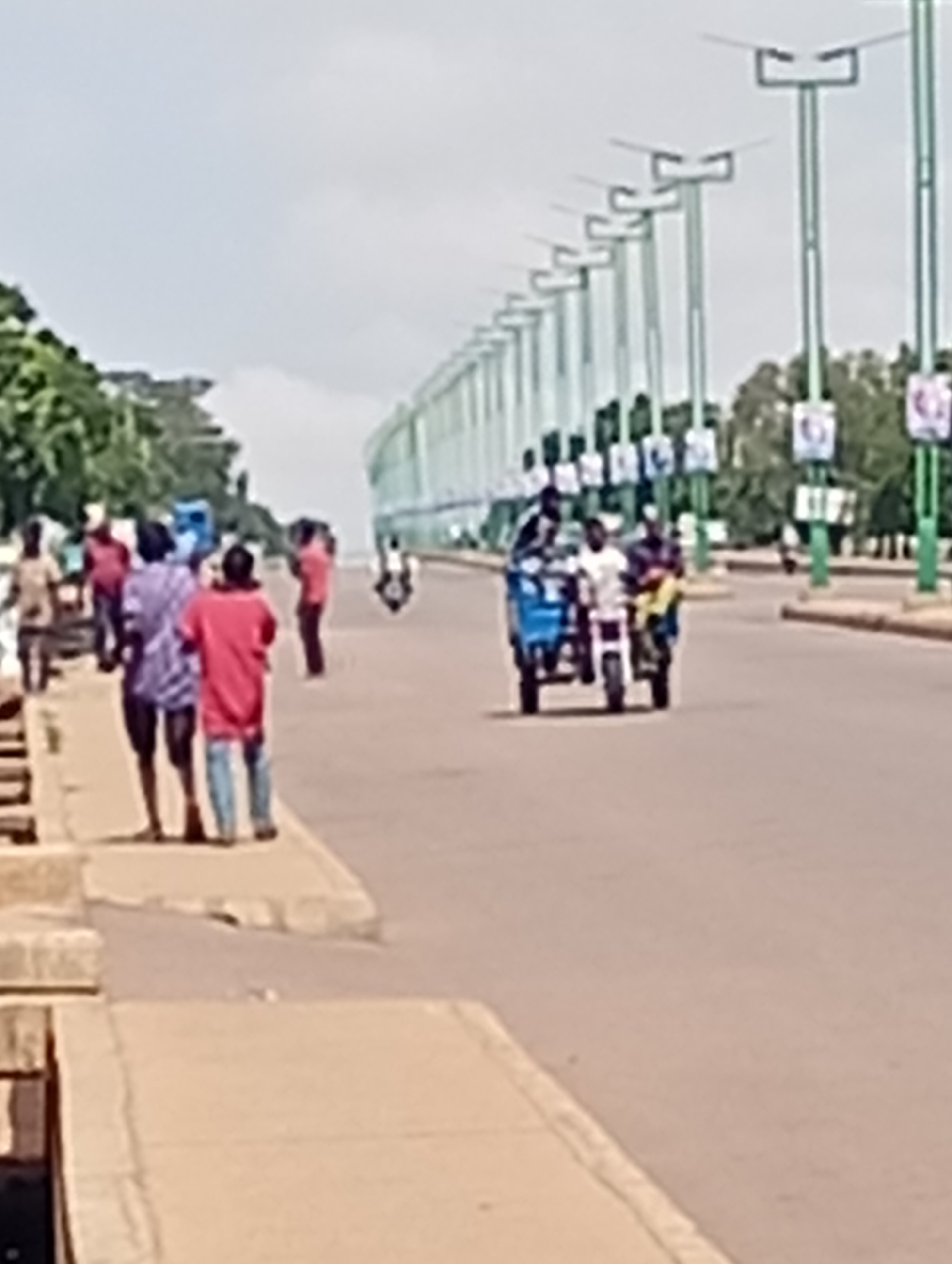 In Tumfure, Nigeria, a street scene unfolds under a hazy sky. A row of tall, green streetlights with horizontal fixtures lines the right side of a paved road. Billboards displaying images of people are attached to some of these lights. On the road, a blue tricycle with a white front panel is being ridden by three individuals, with another person on a motorcycle behind it. Several people are standing on a paved sidewalk on the left side of the road, some observing the traffic. Trees and foliage are visible in the background, suggesting an urban or semi-urban environment. The overall impression is one of everyday street life.