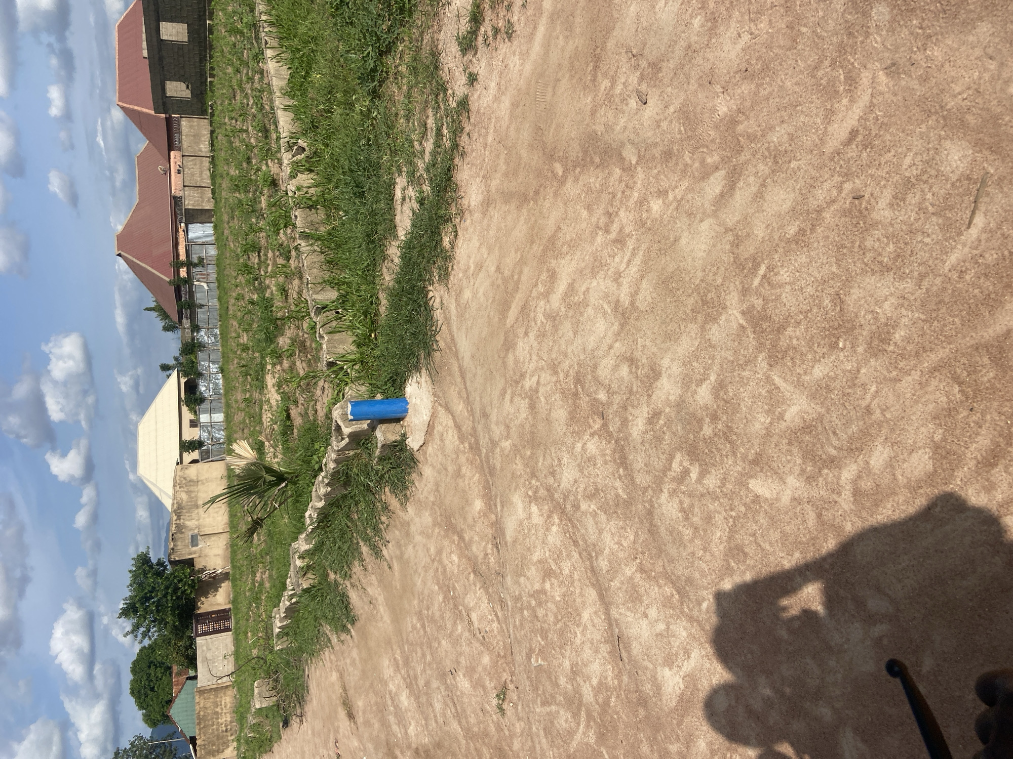 This image, taken in Kuson, Nigeria, depicts an outdoor scene with a construction site and a dusty ground. The foreground is dominated by dry, reddish-brown earth, with tire tracks and some sparse vegetation visible. On the left side of the frame, there are buildings under construction. One building has a red, pitched roof and appears to be made of concrete blocks. Another building, lighter in color, also has a pitched roof and some windows.
A prominent object in the middle ground is a blue cylindrical object, possibly a pipe or a marker, placed on a concrete structure that is partly covered by grass. Beyond these buildings, more structures and trees can be seen, indicating a developing area. The sky is visible in the upper left, with fluffy white clouds against a blue background, suggesting it is daytime with fair weather.
The image has a vertical orientation, likely captured by a mobile phone. A shadow of a person or an object, possibly a bicycle or motorcycle due to the shape of the handlebars visible in the bottom right, is cast on the ground. The overall impression is one of ongoing development in a semi-arid or dusty environment. No visible text is present in the image.