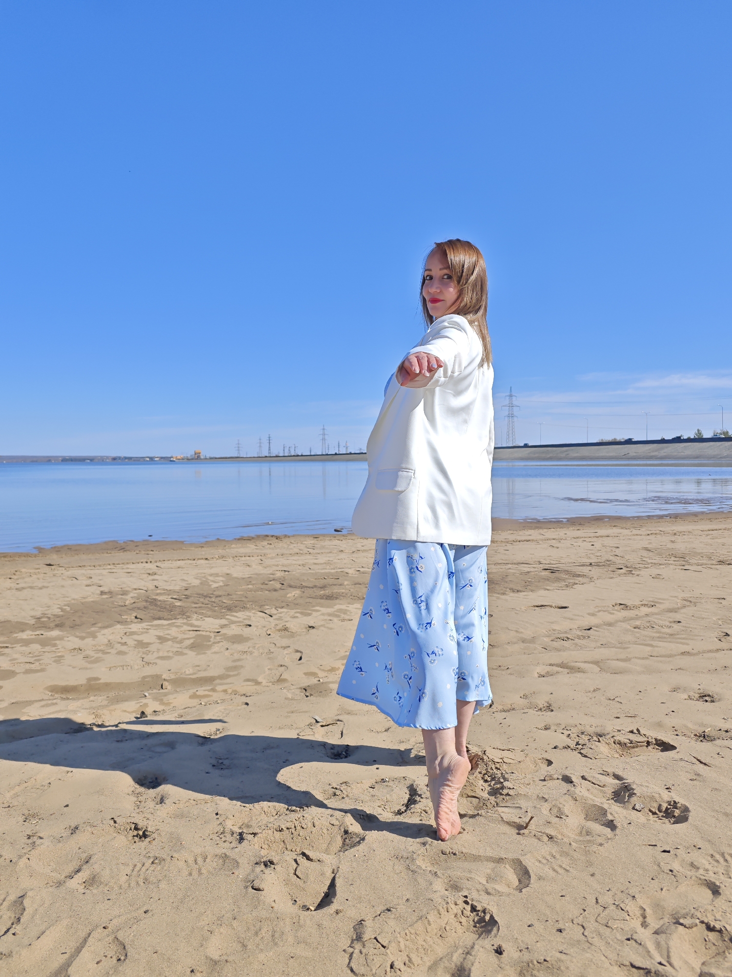 A woman stands on a sandy beach next to a body of water under a clear blue sky. She is wearing a light blue dress with a floral pattern, a white blazer, and is barefoot. She is facing away from the camera but is turned slightly towards the viewer, looking back over her shoulder. The beach has visible footprints and shadows. In the background, across the water, is a landscape that includes power lines, industrial structures, and what appears to be a highway with cars. The location is Noviy, Russia.