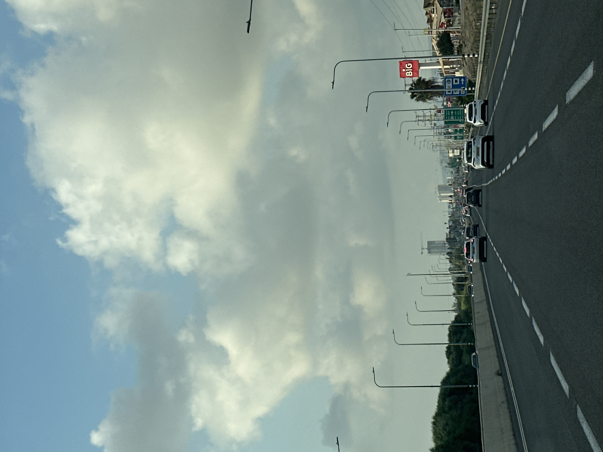 The image depicts a view from a vehicle traveling on a multi-lane highway. The road surface is asphalt, with clear white dashed lane markings. On the left side of the highway, there is a concrete barrier followed by a dense line of green trees. On the right side, there are numerous streetlights, power lines, road signs, and some buildings. One prominent red sign displays the word "BIG". In the distance, several tall buildings and construction cranes are visible, suggesting an urban or suburban area. The sky is partly cloudy with large, fluffy white clouds against a light blue background. Several cars are on the road, moving in the same direction as the viewer's vehicle. The scene appears to be captured during daylight, possibly late afternoon or early morning due to the lighting. The location is identified as the Mate Asher Regional Council in Israel.