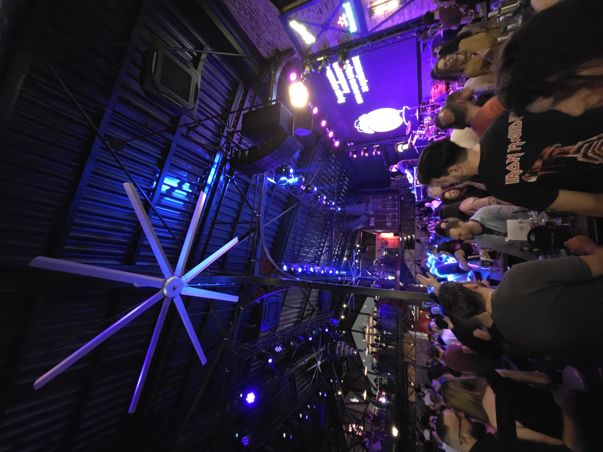The image shows an interior shot of a crowded venue in Varna, Bulgaria. The space is dimly lit with blue and purple lighting, which appears to be stage lighting, as there is a stage set up in the background, with a band playing on it. Patrons stand or sit at tables and along the bar, many of them facing the band. Some are wearing t-shirts with band logos, including one that reads "Iron Maiden". The architecture of the venue features an exposed metal ceiling with visible ductwork and support beams. A large, non-rotating, white ceiling fan is prominently positioned in the left part of the frame. The overall atmosphere is lively, consistent with a live music performance.