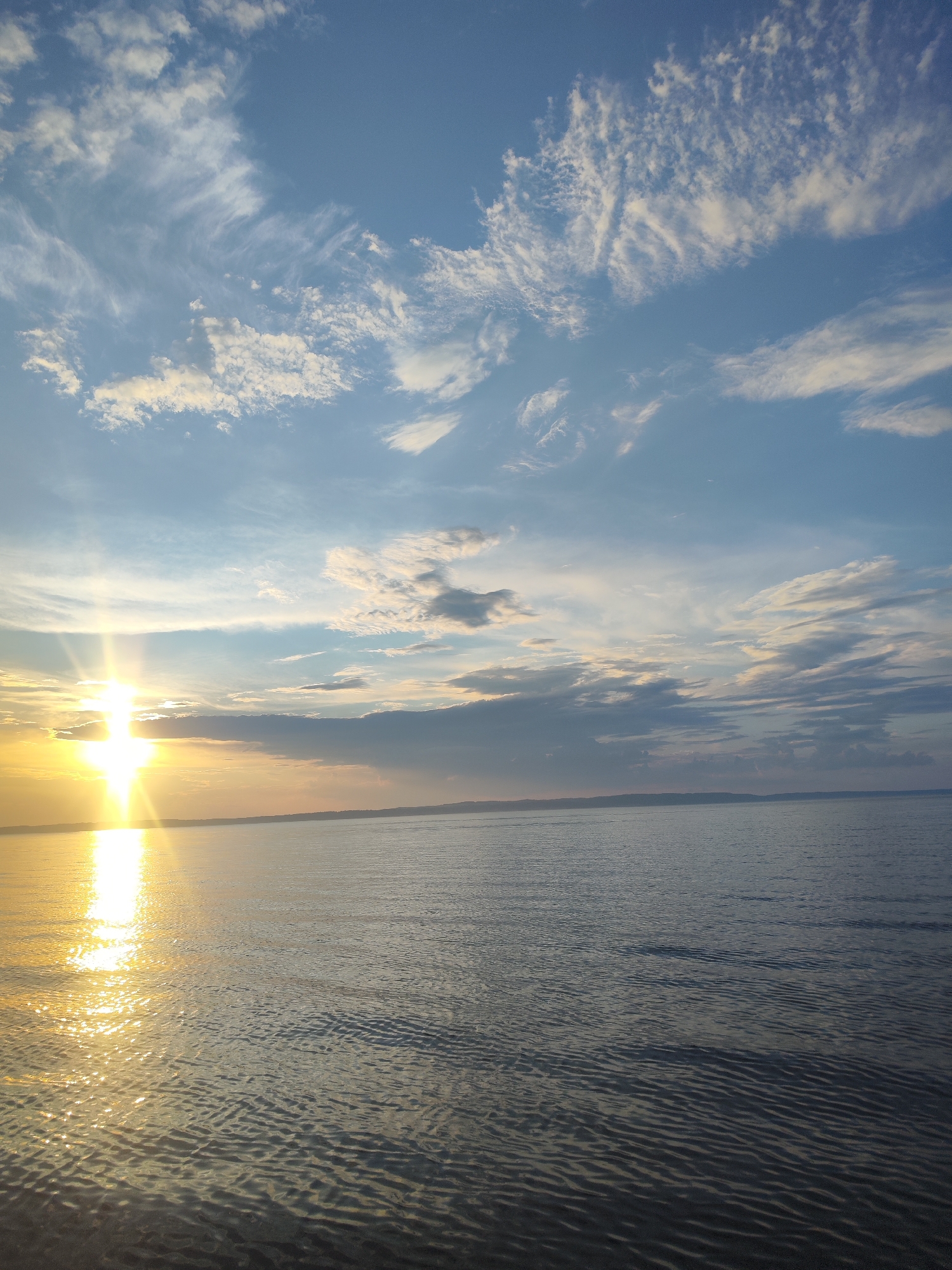 This image depicts a scenic view of a body of water under a sky with scattered clouds and a bright sun. The sun, positioned on the left side of the frame, casts a brilliant golden reflection on the rippling surface of the water. The sky transitions from a bright blue near the sun to a softer hue further away, with wispy white clouds scattered across it. A distant horizon line suggests the presence of land. The overall impression is one of a tranquil natural landscape, likely a lake or sea. The photograph was taken in the city of Niva, Russia.
