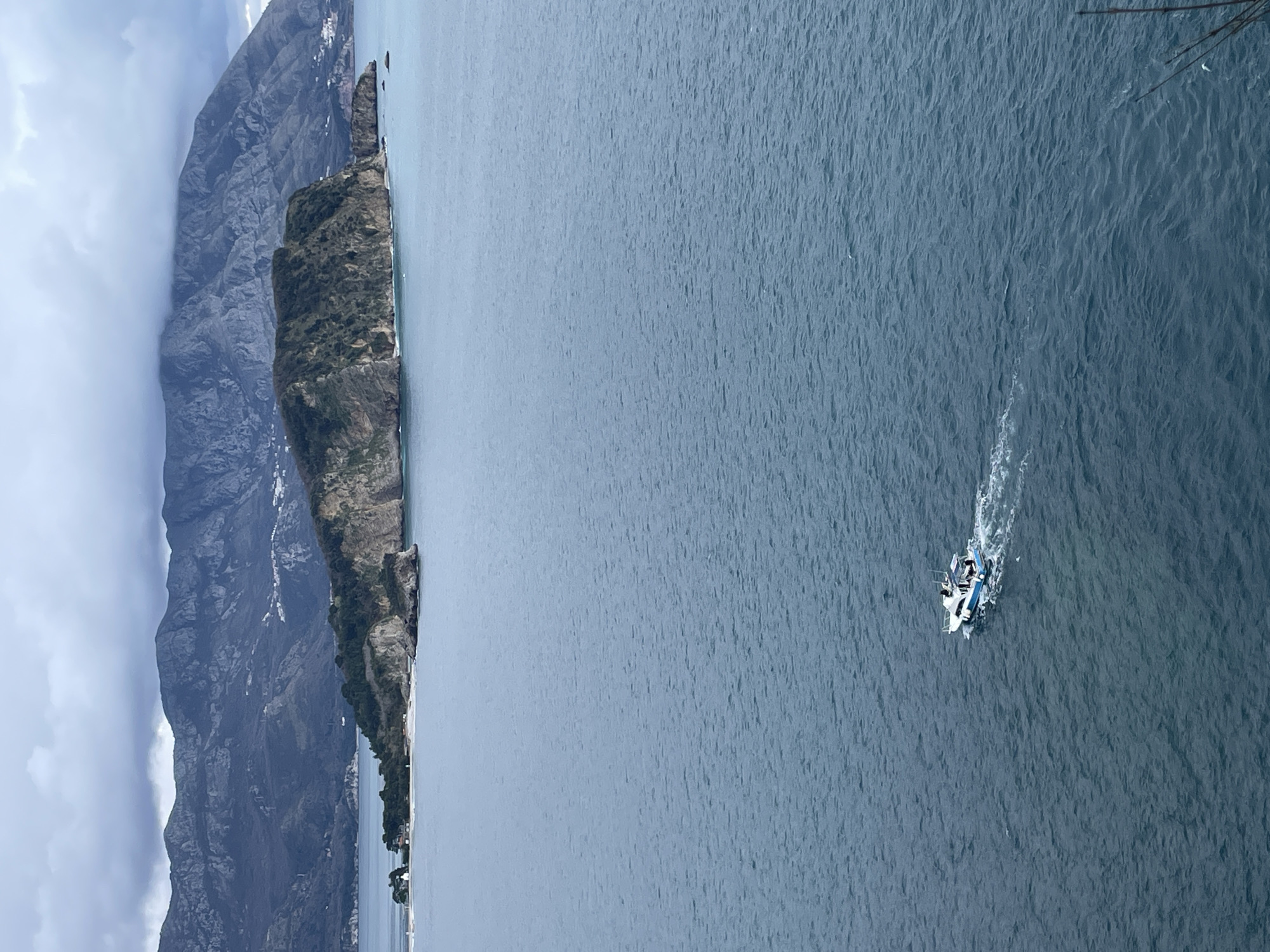 The image displays a coastal panorama from an elevated perspective in Budva, Montenegro, looking towards Sveti Nikola Island. The foreground consists of dark blue-grey ocean water, exhibiting small surface ripples. A white and blue motorboat, carrying at least two individuals, is located in the lower-middle right, actively moving and generating a distinct white wake. In the mid-ground, Sveti Nikola Island (St. Nicholas Island) is visible, characterized by rugged, vegetated cliffs and rocky formations. A narrow, lighter-colored land spit, featuring a small white building, extends from the left side of the island. The background reveals a range of dark, rugged mountains, partially shrouded by an overcast, grey sky. Scattered white structures indicative of human habitation are visible on the lower slopes of these distant mountains.