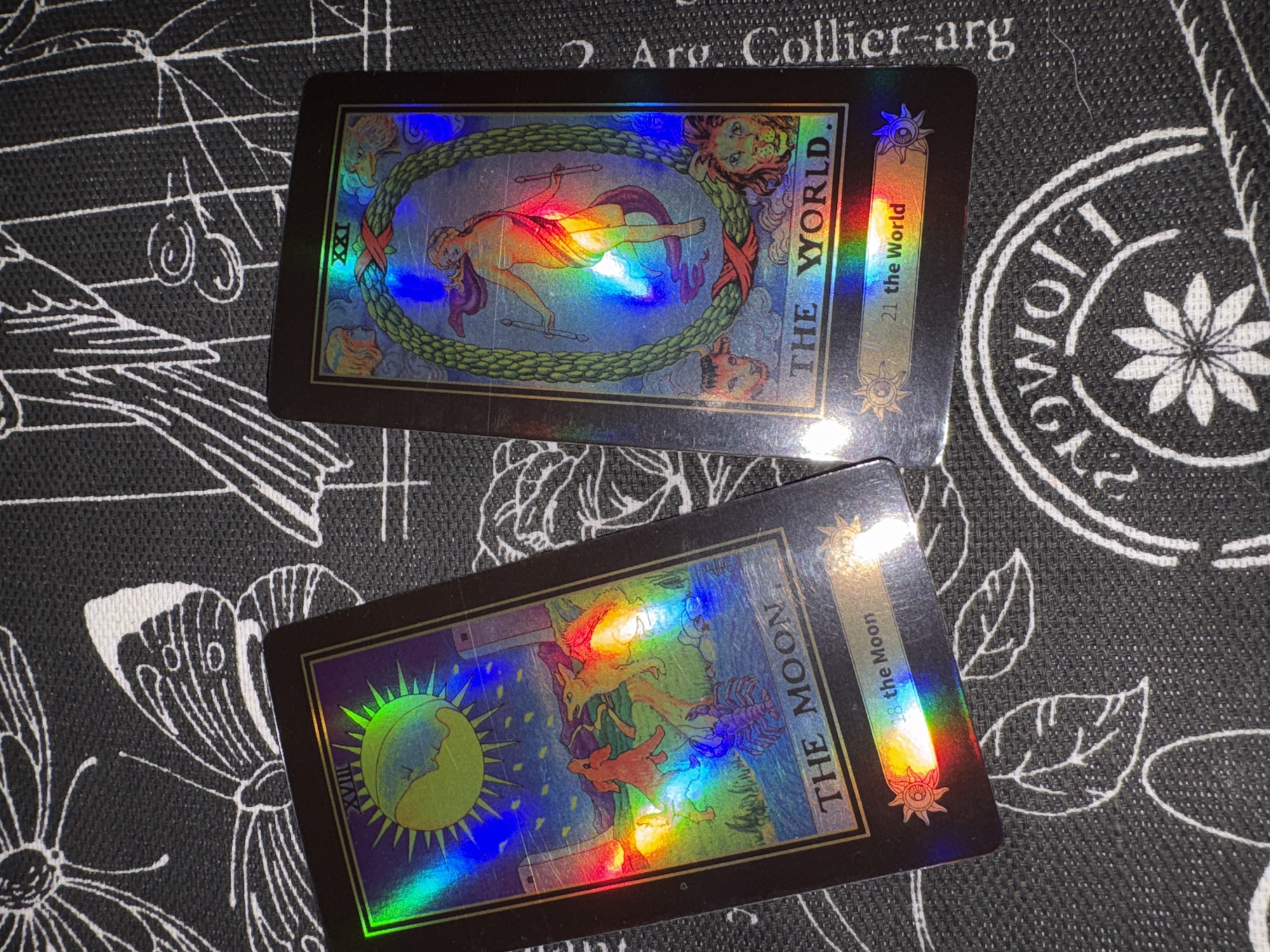 The image displays two holographic tarot cards positioned on a dark, woven fabric surface. The fabric features white line-art patterns, including a butterfly and leaves on the left, and abstract swirls along with an inverted circular motif containing a central multi-petal flower on the right.
The card on the left is "The Moon" (XVIII / 18), oriented slightly diagonally. Its artwork depicts a large crescent moon with a human face radiating spiky light rays against a dark blue sky. Below, two towers flank a body of water from which a red scorpion or lobster emerges. Two canids, one appearing wolf-like and the other fox-like, are depicted howling or looking upwards between the towers and the water. The card's surface exhibits strong rainbow iridescent reflections from a light source. The title "THE MOON" is printed in gold at the bottom.
The card on the right is "The World" (XXI / 21), also slightly diagonal and partially overlapping the left card. It features a central nude human figure, appearing female, draped with purple and red fabric, holding two wands or batons, depicted in a dancing or walking pose within a large green laurel wreath. The wreath is bound with red sashes at the top and bottom. Four symbolic figures—a blonde human head (top left), an eagle's head (top right), a bull's head (bottom left), and a lion's head (bottom right)—are depicted outside the wreath at the corners. This card also exhibits significant rainbow holographic reflections across its imagery. The title "THE WORLD." is printed in gold at the bottom.
Both cards have black borders with a thin inner gold line and feature stylized sunburst emblems at the bottom corners of their informational strip. The lighting creates prominent specular reflections across the holographic elements of both cards. The image provides no identifiable geographic markers for any specific city, including Izhevsk, Russia.