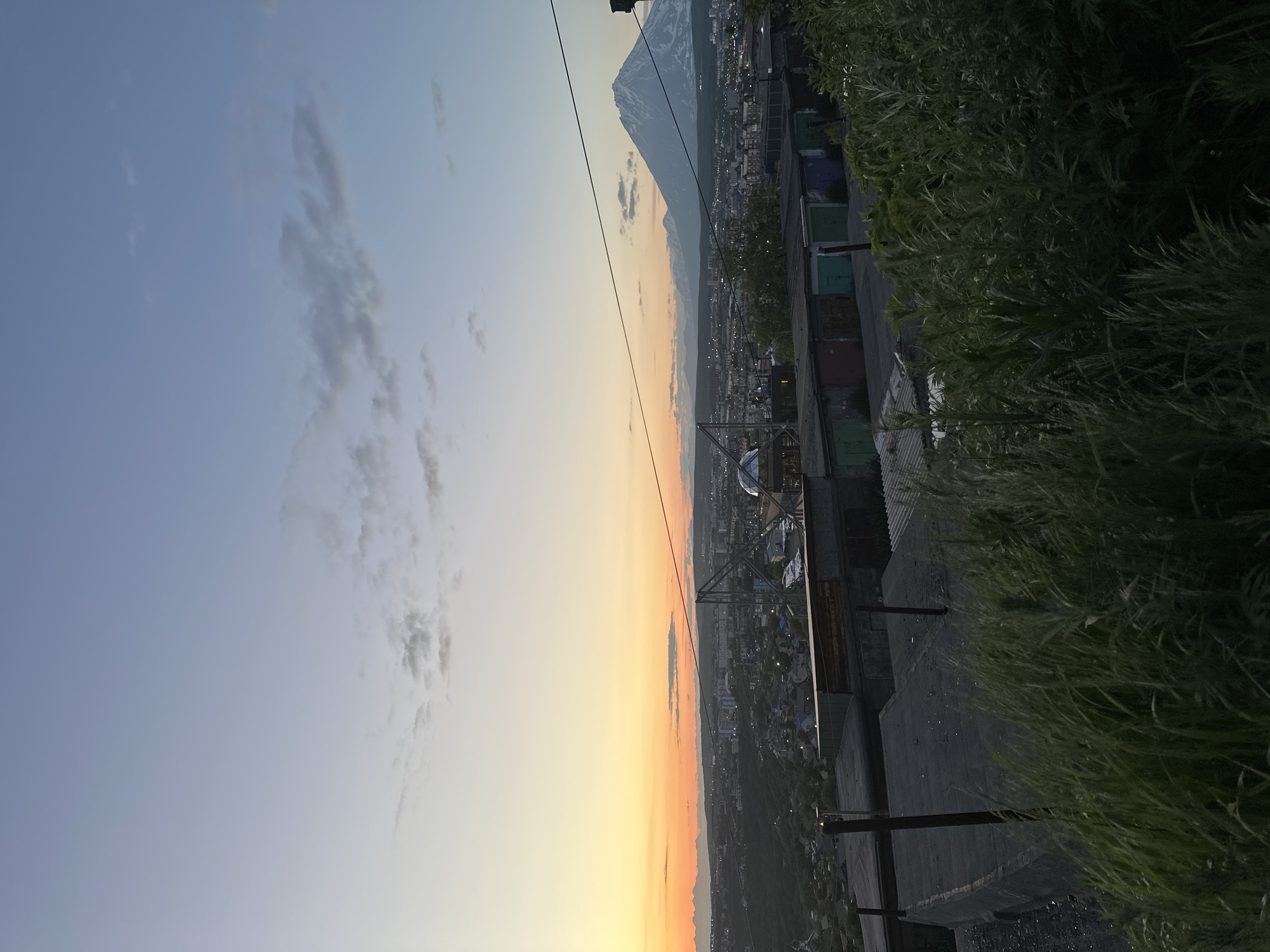The image depicts a panoramic view of the city of Petropavlovsk-Kamchatsky, Russia, at dusk. In the foreground, tall green grass and what appear to be concrete structures or flat roofs are visible. Beyond this, a row of colorful garage doors runs horizontally across the mid-ground, suggesting an urban or suburban area. Further into the distance, the city's buildings are spread out, with lights beginning to appear. Dominating the background is a majestic, snow-capped volcano, identifiable as a prominent landmark of the region. The sky is a gradient of soft blues and oranges, indicative of sunset, with a few scattered clouds. Power lines traverse the frame, connecting different parts of the scene. The overall impression is a serene, twilight landscape showcasing the natural beauty of Kamchatka juxtaposed with urban development.