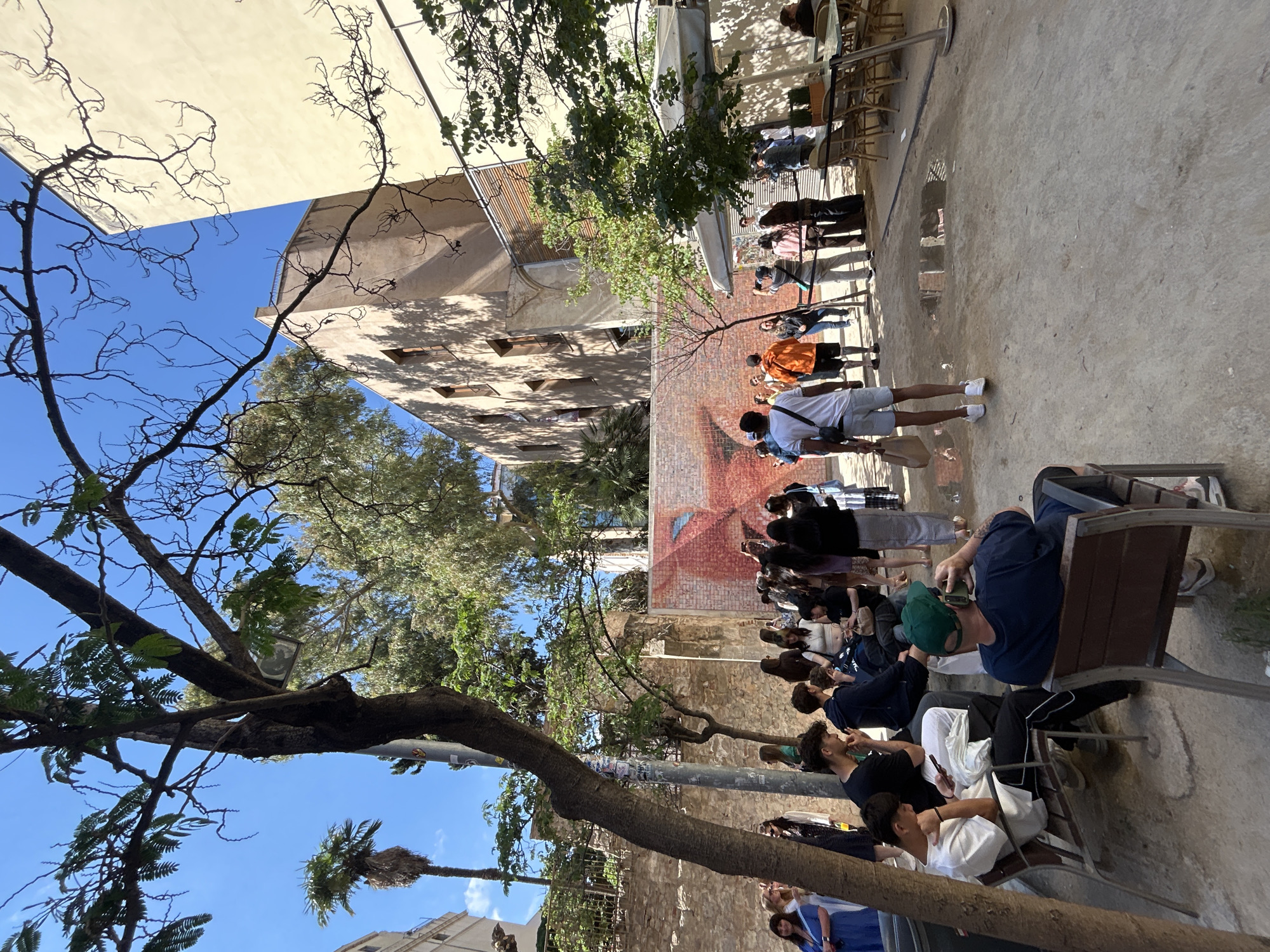 The image depicts a courtyard in Barcelona, Spain, with a crowd of people lining up. Some are seated on benches, while others stand in a queue that stretches towards a building with a brick mural. The courtyard is surrounded by buildings, some with exposed brickwork, and is partly shaded by trees. The sky is blue, suggesting a sunny day. People are wearing casual clothing.