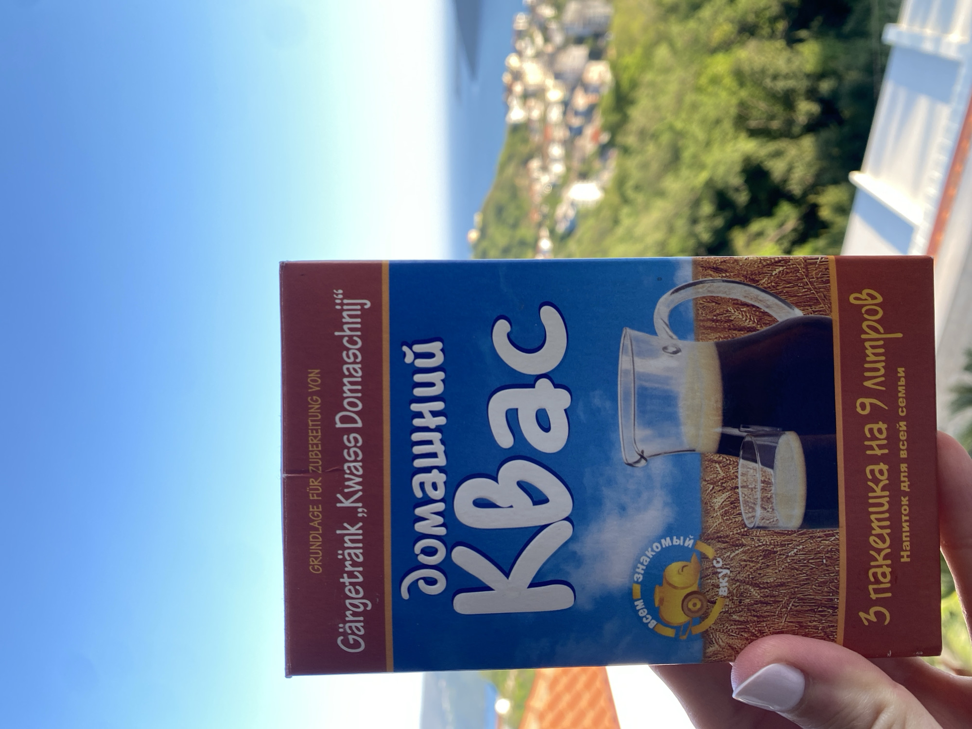 The image captures a hand holding a package of Kvass in an outdoor setting, overlooking a scenic landscape. The dominant subject is a blue and brown rectangular package, held vertically by a hand with light-colored nail polish. The package is for "Kvass Domaschnij" (Homemade Kvass), a traditional fermented drink.
The background reveals a bright, clear blue sky dominating the upper left, while the lower right shows a lush, green, hilly landscape with numerous buildings, suggesting a town or village. Beyond the land, a calm body of water is visible, likely a sea or large lake, stretching into the horizon under the bright daylight. The scene suggests a view from a balcony or terrace, characteristic of a coastal or mountainous region like Trebesin, Montenegro.
Visible text on the package, in a mix of German and Russian, includes:
- "GRUNDLAGE FÜR ZUBEREITUNG VON Gärgetränk „Kwass Domaschnij"" (German: Base for preparation of fermented drink "Kvass Homemade")
- "домашний Квас" (Russian: Homemade Kvass)
- A circular logo with a bird and text "всем знакомый вкус" (Russian: familiar taste to everyone).
- "3 пакетика на 9 литров" (Russian: 3 sachets for 9 liters)
- "Напиток для всей семьи" (Russian: Drink for the whole family)
An illustration of a pitcher filled with Kvass and a glass is also prominent on the package. The weather is clear and sunny, indicating daytime.