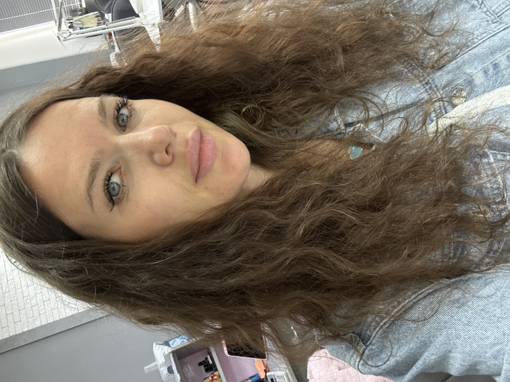 The image is a portrait of a young woman, taken indoors and rotated approximately 90 degrees counter-clockwise. The woman, with her head tilted to the left, gazes directly at the camera with a calm, neutral expression. She has long, wavy, rich brown hair that cascades around her face and shoulders. Her eyes are a striking blue-grey, framed by prominent, dark eyelashes. Her lips are full and have a natural, light pink hue.
She is wearing a light blue denim jacket, and a small, light blue or teal pendant is subtly visible on a chain near her neck, partially nestled within her hair. The background consists of an indoor setting, with bright, even lighting. To her upper left, a textured wall with a white rectangular tile or brick pattern is visible. Below this, a smooth, light grey surface occupies a significant portion of the background. Further back, indistinct objects include what appears to be a black chair and a metallic rack with white components. Other blurred shapes, including a pink item, can be discerned in the lower left area of the image. The scene suggests a modern interior, possibly a salon or a contemporary living space, located in Moscow, Russia. No visible text is present in the image.