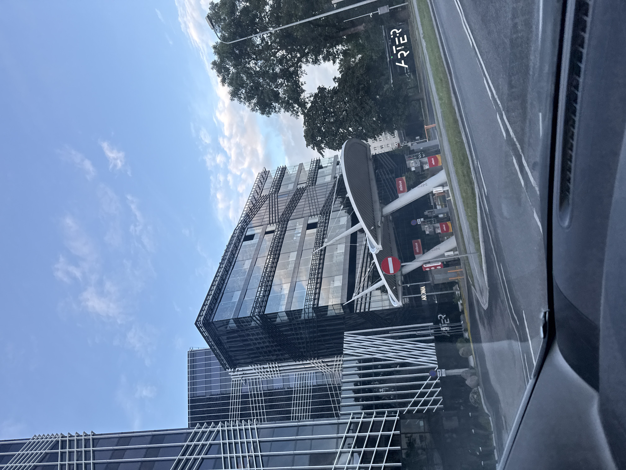 A modern architectural scene unfolds in Tallinn, Estonia, captured from a low vantage point, likely from within a vehicle. Dominating the left side of the frame is a tall, sleek glass skyscraper with a distinctive white lattice facade. Adjacent to it, another contemporary building features a dark, reflective glass exterior. In the mid-ground, a distinctive, futuristic-looking gas station canopy with white supports and a curved roof stands prominently. Red and white signage, along with red fuel pumps, are visible beneath the canopy. The words "AROMA" and "miles+" are discernible on some of the station's elements. To the right, mature trees with dark green foliage provide a natural contrast to the urban structures. The sky above is a pale blue, punctuated by scattered white clouds. The road in the foreground is asphalt, marked with white lines, indicating a street or highway. A black car interior, including a dashboard section, is visible at the very bottom, reinforcing the perspective of being inside a vehicle. The overall impression is one of a contemporary cityscape with a focus on modern architecture and infrastructure.