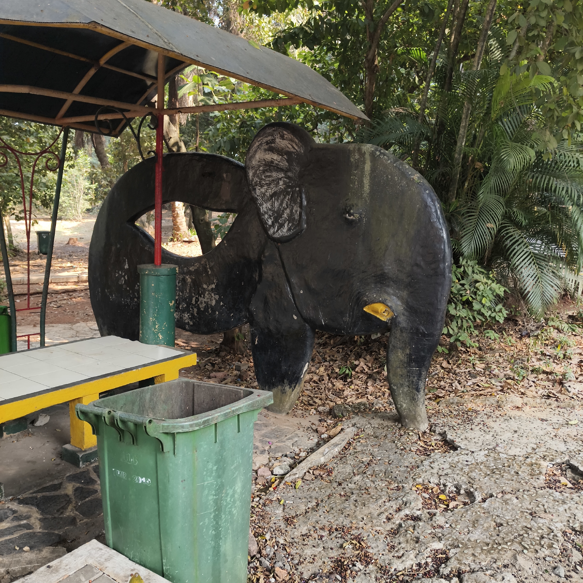 The image depicts an outdoor scene, likely a park or recreational area, with a sculpture of a dark-colored elephant prominently featured. The elephant sculpture is black and appears weathered, with some lighter patches of what might be paint or material showing through. It has large ears and a visible tusk area, with a yellow piece attached to what could be its mouth. The sculpture stands on a ground covered with fallen leaves and a rough, pebbly surface.
To the left of the elephant, there is a picnic table with a yellow frame and a white tiled surface. In front of the table, a large, green plastic trash bin is visible. Behind the elephant and to the right, lush green foliage and trees form a dense background. A portion of a rustic metal canopy structure with a dark roof is visible in the upper left quadrant of the image, supported by wooden beams and metal poles. The overall lighting suggests it is daytime.
The image context provided indicates the location as the city of South Jakarta, Indonesia. This specific scene appears to be within Taman Pintar Jakarta, a park in South Jakarta. The presence of the elephant sculpture, picnic table, and trash bin are typical features found in such public spaces, often designed for family recreation and education. The natural environment with trees and greenery points to a well-maintained outdoor area.