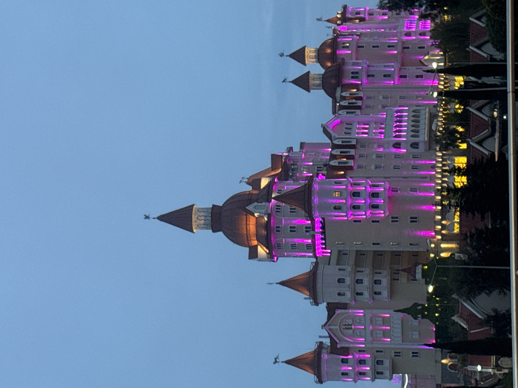 The image shows a building resembling a castle illuminated with purple and yellow lights at night. The building has multiple towers with conical roofs and decorative elements. The sky is a dusky blue, suggesting either late evening or early morning. In the city of Sirius, Russia, this building may be associated with a tourist attraction, such as the Sochi Park theme park.