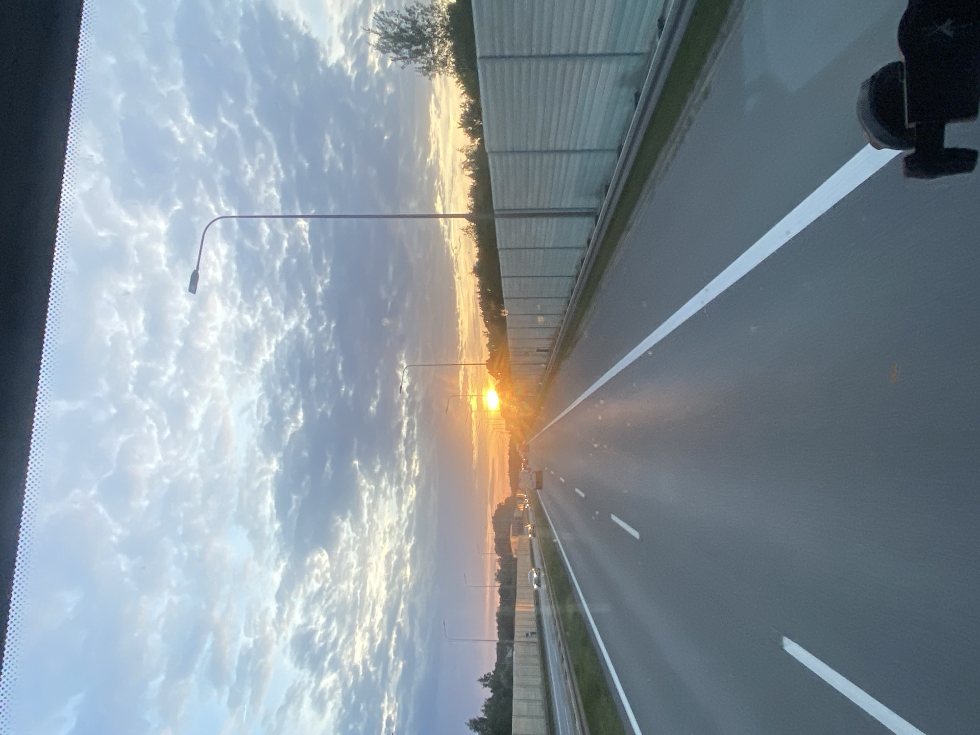 The image depicts a highway scene at sunset in Podłęże, Poland. The perspective is from inside a vehicle, looking forward. The road is paved with asphalt and has clear white lane markings. On the right side of the road, there is a tall, light-colored sound barrier. On the left side, there is a similar sound barrier and vegetation. Several cars are visible on the highway, moving in the same direction as the viewpoint. The sky is filled with dramatic clouds, tinged with the orange and yellow hues of the setting sun, which is prominently visible on the horizon, casting a bright glow and reflections on the road. Streetlights are also present along the highway. The overall impression is of a journey through a rural or suburban area during a beautiful sunset.