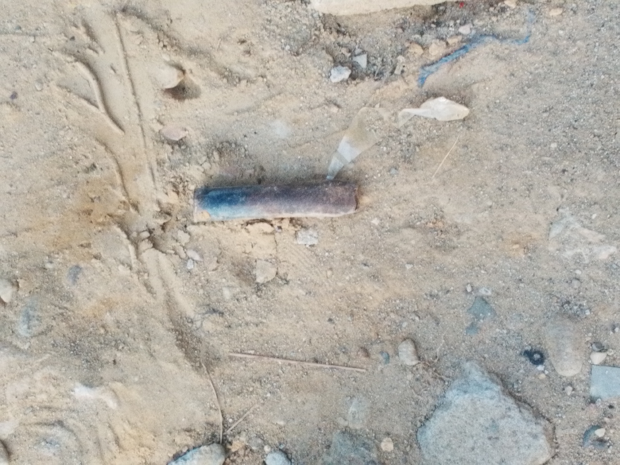 The image depicts a patch of sandy, gravelly ground in an outdoor setting. At the center is a cylindrical object with dark coloration at one end transitioning to a rusty brown hue. Debris such as small rocks, plastic pieces, and a thin twig are scattered throughout. The sandy surface exhibits textural variation and minor indentations, including what appears to be a footprint or a marking. The overall environment suggests an unkempt or disturbed area. There are no people present. The location in the city of Lhokseumawe, Indonesia, is not discernible from the image alone, and no contextual information is available to confirm that it is the location of this image.