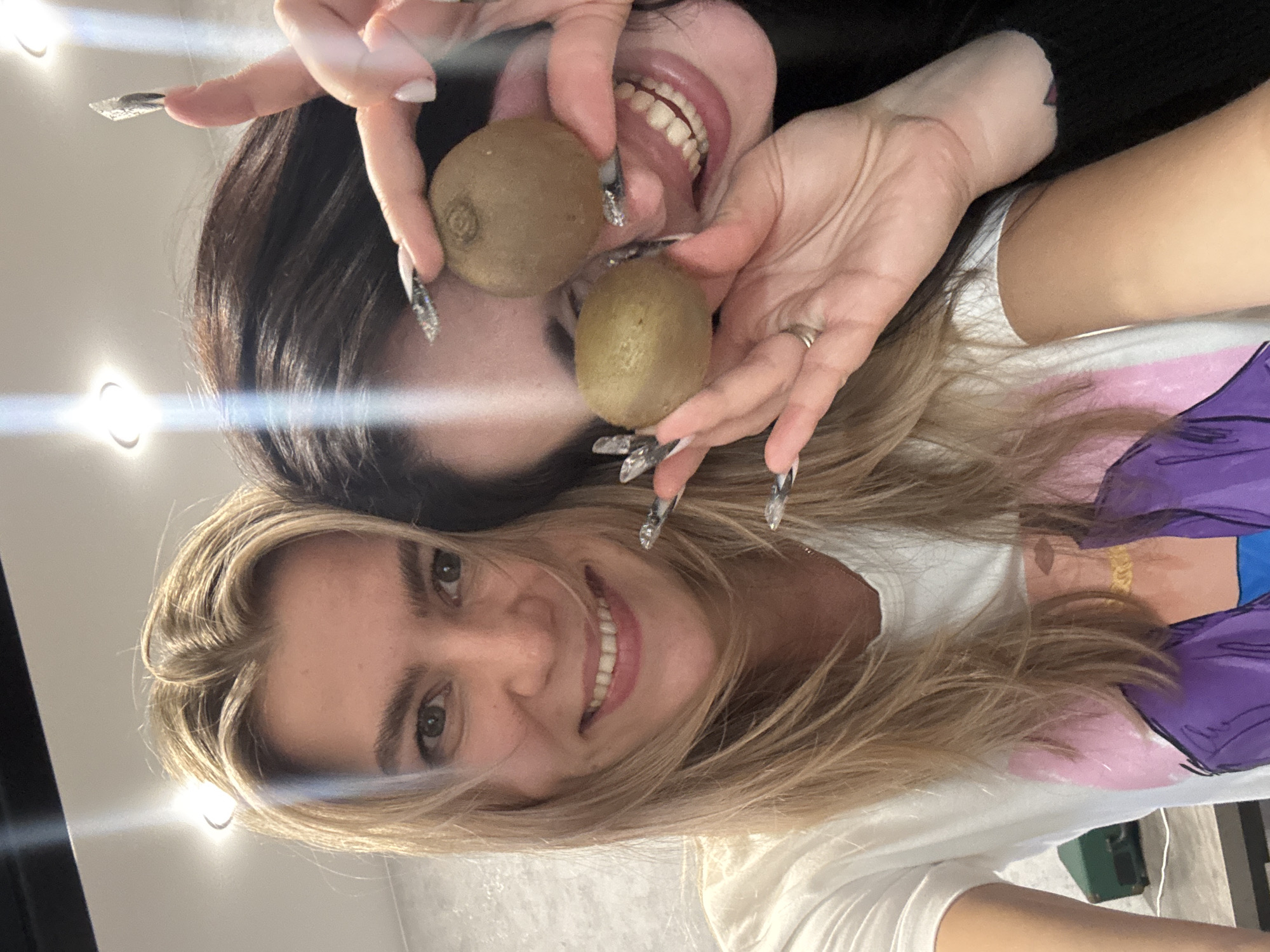 This image captures a playful moment between two women, taken from an overhead perspective. The woman in the upper portion of the frame has long dark hair and is smiling widely with her mouth open. She holds two kiwi fruits on either side of her mouth, creating a humorous expression. Her long fingernails are prominently visible, adorned with intricate, sparkly designs.
Beneath her, and slightly overlapping, is another woman with long blonde hair, also smiling broadly and looking directly at the camera. She wears a white t-shirt with a colorful graphic design featuring elements in purple and pink. A ring is visible on her finger.
The background is a plain, light-colored surface, possibly a wall or ceiling, featuring several bright, circular light reflections, suggesting an indoor setting with artificial lighting. Both women appear happy and engaged in the playful pose. No visible text is present in the image.