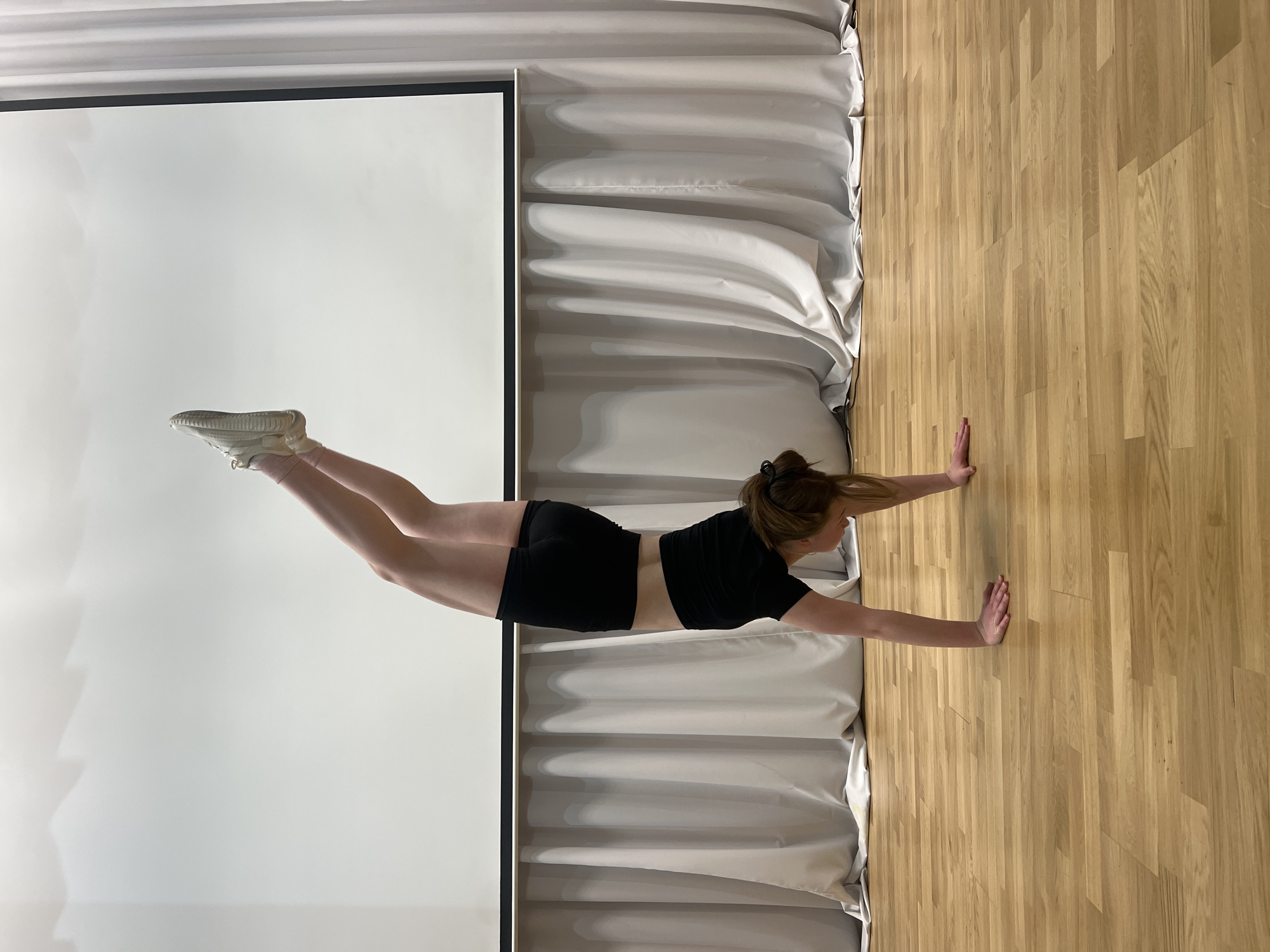 A young woman is positioned horizontally in a room. Her hands are pressed against a wood-paneled wall to her right. She is wearing a black crop top and black shorts, with white shoes on her feet, which are pointed toward a white screen to her left. Her body is in a straight line. The room has white curtains behind the woman, positioned between the wall and the screen. The location may be Chaikovsky, Russia, though there is no specific evidence of this in the image.