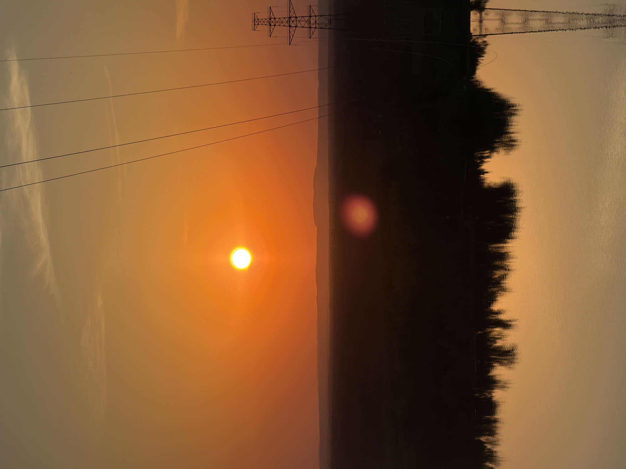 The image shows a sunset with a partially visible sun against an orange sky. Power lines and a power tower are silhouetted against the sky on the upper left. A dark horizontal mass stretches across the middle of the image, likely representing land or trees. Its reflection is visible in the water on the right. The location is likely Noviy, Russia. There are no people visible.