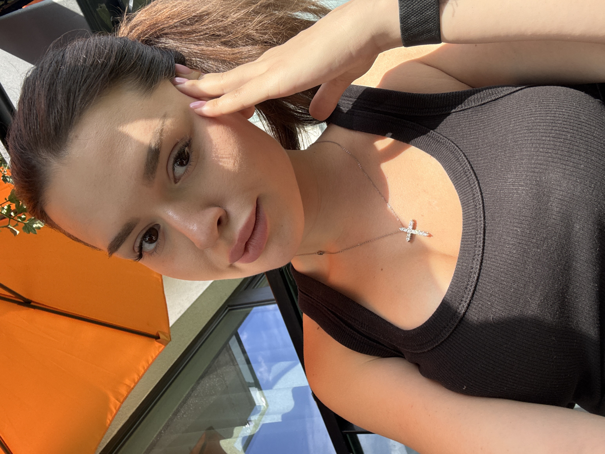 A woman with brown hair in a ponytail, wearing a black tank top and a silver cross necklace, takes a selfie outdoors. She is leaning her head on her hand, her right arm resting on her chest. Sunlight casts shadows across her face and shoulders. In the background, an orange umbrella is visible, along with a glass window reflecting a blue sky with clouds.