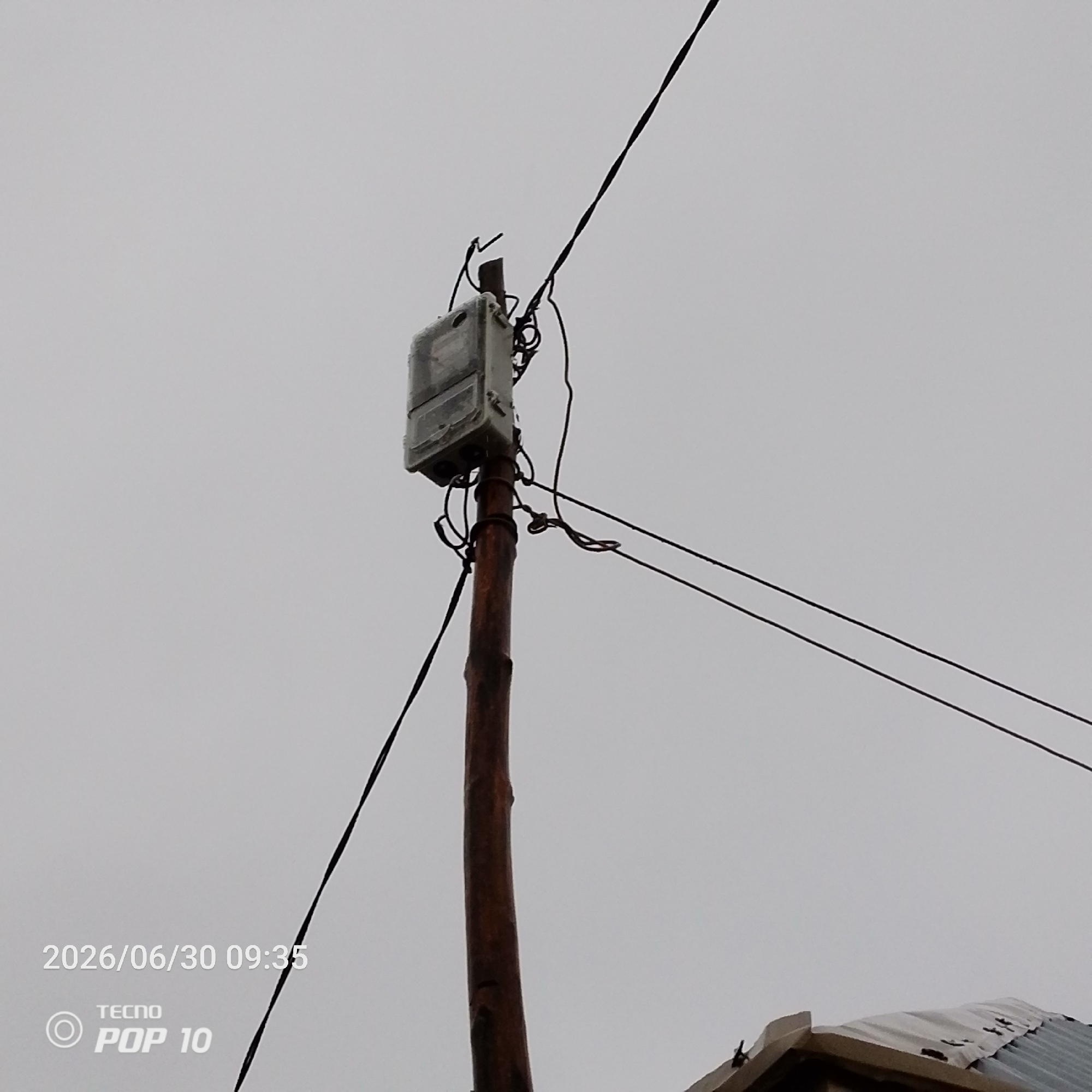 A power meter is mounted on a wooden utility pole, attached with wires. The meter is in a weathered grey casing, and the pole is dark brown and appears rough. Electrical wires are strung across the frame, some taut and others slack. In the lower left corner, a timestamp reads "2026/06/30 09:35" along with the TECNO POP 10 logo. In the bottom right corner, a section of a corrugated metal roof is visible. The sky is overcast and light grey. The image appears to have been taken outdoors in an urban or semi-urban environment, likely in Jalingo, Nigeria, given the timestamp and branding that suggest a mobile phone used for photography.