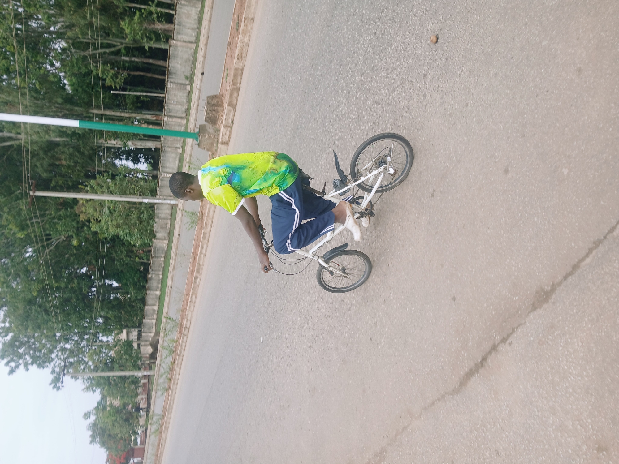 A man rides a white BMX bike on a paved road in Bukuru, Nigeria. He wears a green and yellow t-shirt and navy blue track pants with white stripes. The road is cracked in one area. Trees and a green and white pole line the side of the road.