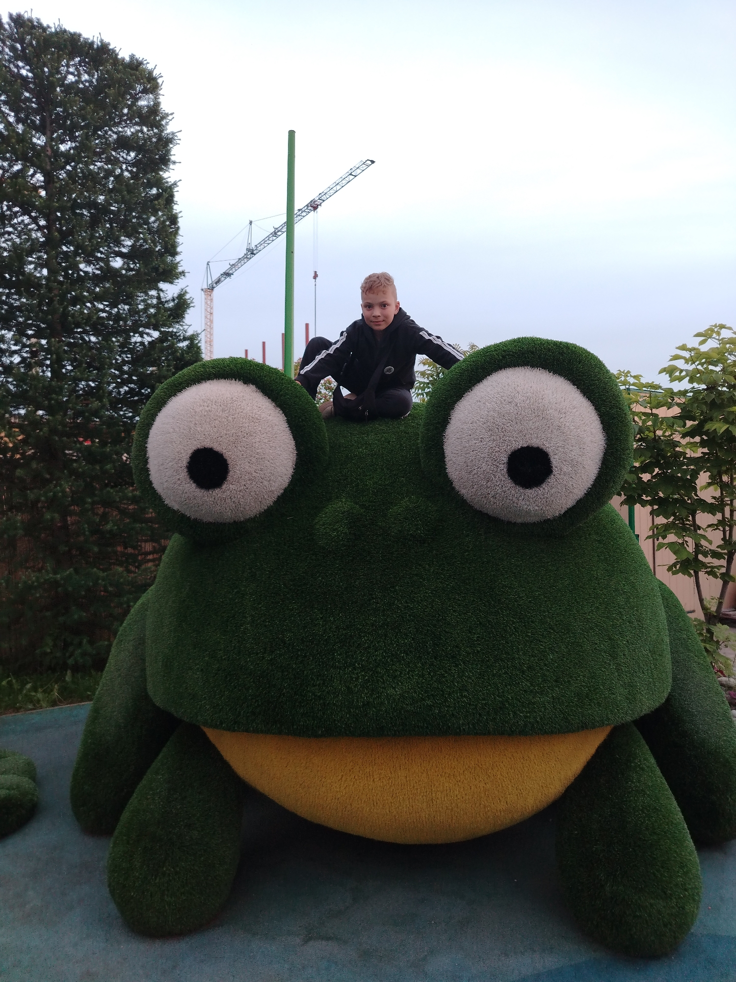 A full shot captures a young person sitting atop a large, artificial frog. The frog is primarily green with a yellow belly and oversized, white eyes with black pupils. The child is wearing a dark-colored jacket with white stripes on the sleeves and dark pants, sitting cross-legged with their arms outstretched to the sides.
The background reveals a setting with trees, a green pole, and two construction cranes.
The image was taken in the city of Kazan, Russia.