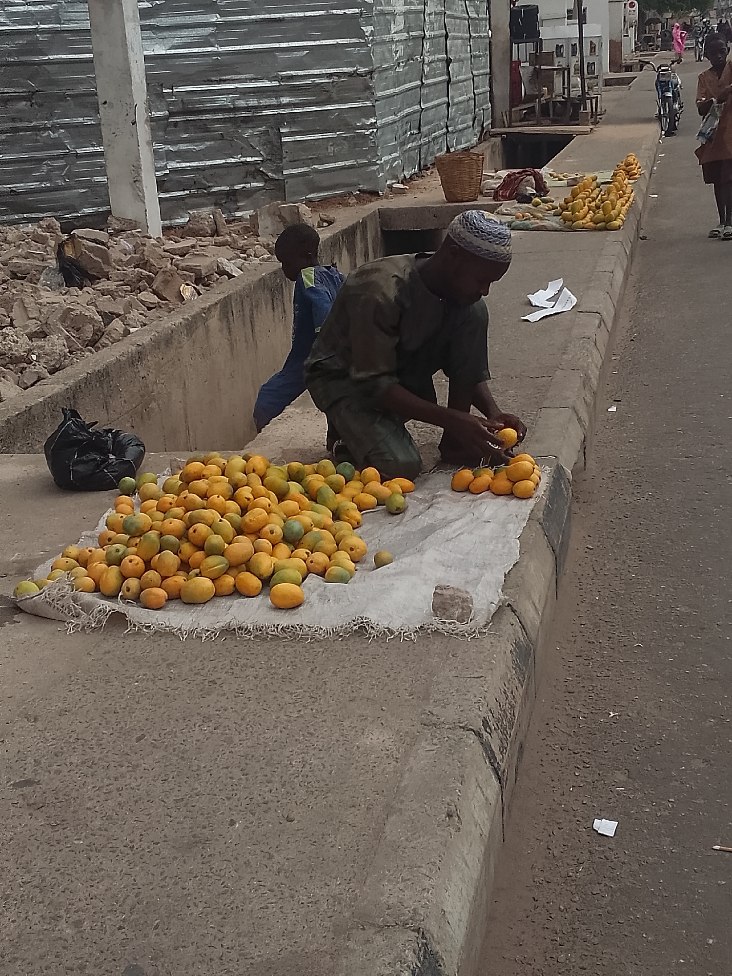 A man is sorting mangoes on the side of a street in Pantame, Nigeria. He is kneeling next to a pile of mangoes that are spread out on a white cloth. He is wearing a blue and white skullcap, and a dark green tunic. Another boy is standing next to him. To the left of the mangoes is a black trash bag, and to the right are more mangoes lined up along the curb. In the background, there are buildings made of corrugated metal, and people walking down the street.