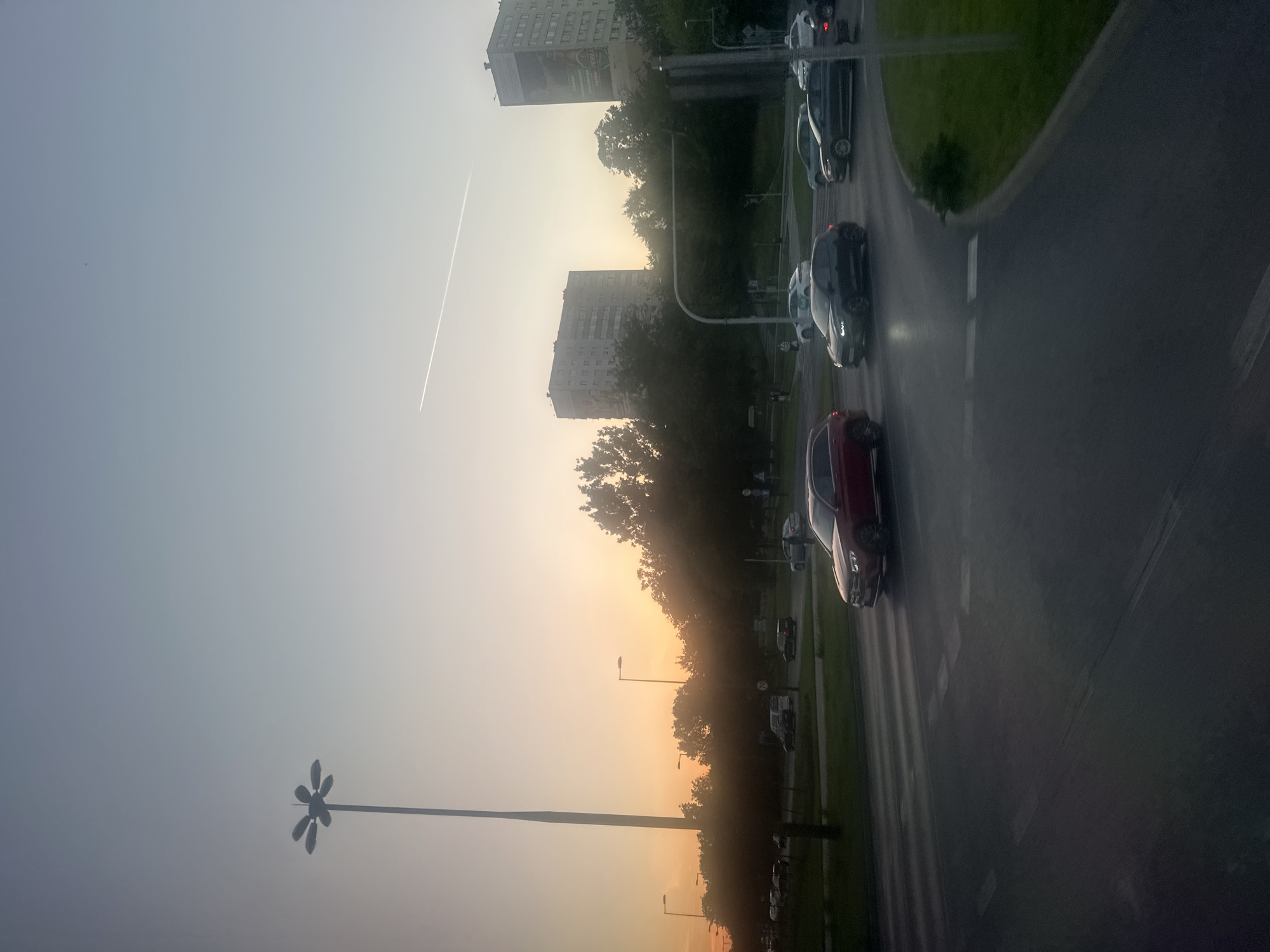 The image depicts a street scene in Lublin, Poland, at dusk. A tall lamppost with multiple arms stands prominently on the left side of the frame. In the background, two apartment buildings are visible, one taller than the other, with a billboard on the side of the taller building. The sky transitions from a soft orange hue near the horizon to a pale blue overhead, with a faint airplane contrail stretching across the upper portion of the image. Trees line the horizon behind the street. Several cars are in motion on the multi-lane road, including a red SUV, a dark hatchback, and other vehicles further in the distance. Pedestrians are also present, though their figures are small and indistinct. The road surface shows reflections from the streetlights and surrounding environment. The overall atmosphere is that of a calm evening in an urban setting.