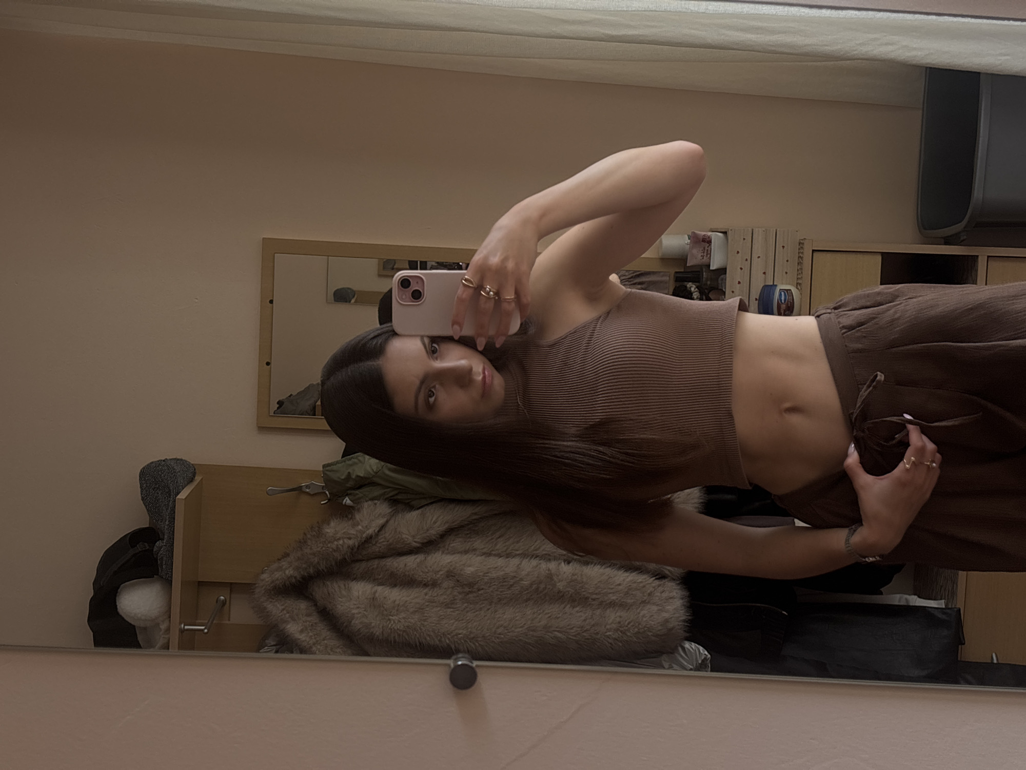 A young woman is taking a mirror selfie in what appears to be a hallway or dressing area. She is standing in front of a large mirror, holding a pink smartphone in her right hand to capture her reflection. She is wearing a brown ribbed crop top that exposes her midriff and matching brown wide-leg pants with a drawstring. Several rings are visible on her right hand.
Behind her, the mirror reflects a hallway with coats hanging on hooks, a shelf with various items including hats and possibly gloves, and a smaller mirror on the wall. To the right of the frame, a sheer white curtain hangs near a window, and a gray trash can is visible. The walls are painted a neutral, light pink color. The overall impression is of a casual, domestic setting. The image does not contain any specific geographical markers to indicate it is located in the city of Varna, Bulgaria.