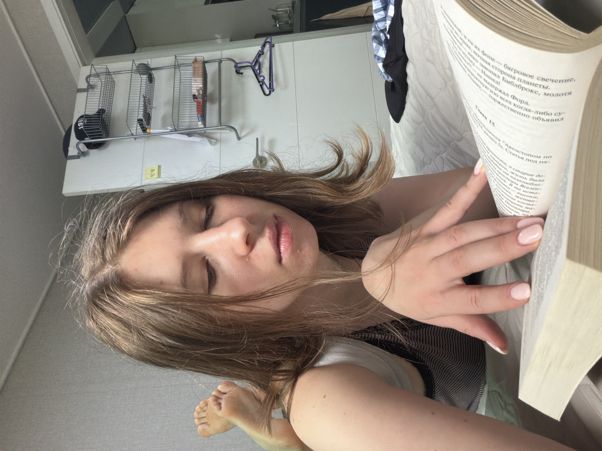 In a residential setting in Dongducheon-si, South Korea, a young woman with light brown hair is lying on a bed, reading a book. Her feet are visible, crossed and resting on the bed. She is wearing a striped tank top. A shelf unit with various items, including baskets and a hat, is attached to a white wall behind her. A clothes rack with hangers is also visible on the wall. The room appears to be a bedroom, with a quilted bedspread and what looks like a closet door in the background.