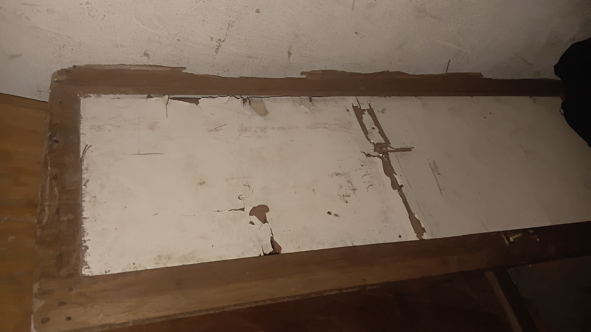 A vertical, full shot of a damaged, off-white wooden doorframe, is pictured at a slight angle to the left. The frame is intact on the left, but on the right, it's peeling away from the wall. The door itself has been removed or broken, leaving only the frame. The interior of the frame is off-white and heavily cracked and peeling. The exterior of the frame is a lighter brown wood. The background is a plain, textured wall, also off-white. The floor is a dark, indeterminate color and appears to be covered by a black cloth. The image is taken in a dimly lit room, likely in Prabumulih, Indonesia, given the context.