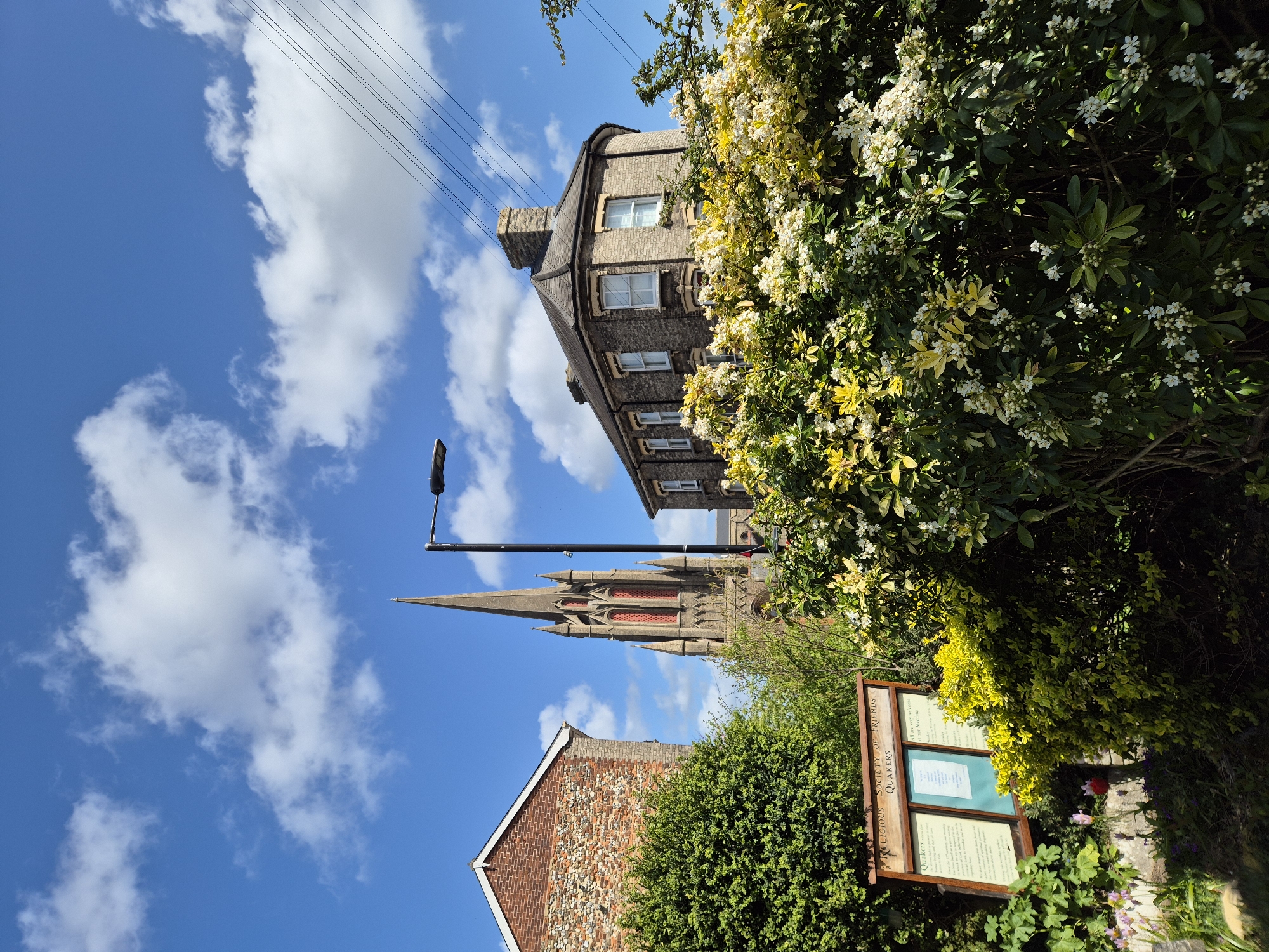 Captured at an oblique angle under a vibrant blue sky dotted with fluffy white clouds in West Suffolk, United Kingdom, the photograph showcases an assortment of architectural styles intertwined with verdant plant life.
Dominating the background is the sharp spire of a church, partly obscured by a light pole. Adjacent to the church, a brown brick building with multiple windows and a unique curved roof stands. In the foreground, a portion of another brick building with a gable roof is visible, its walls a mix of brick and stone.
Lush greenery, including bushes and flowering plants, frame the right side of the image. Among the foliage, a sign for the "Religious Society of Friends Quakers" is partially visible, indicating a place of worship and community.
The clear sky and varied architectural details suggest a sunny day in a historical area within West Suffolk. No people are visible in the scene.