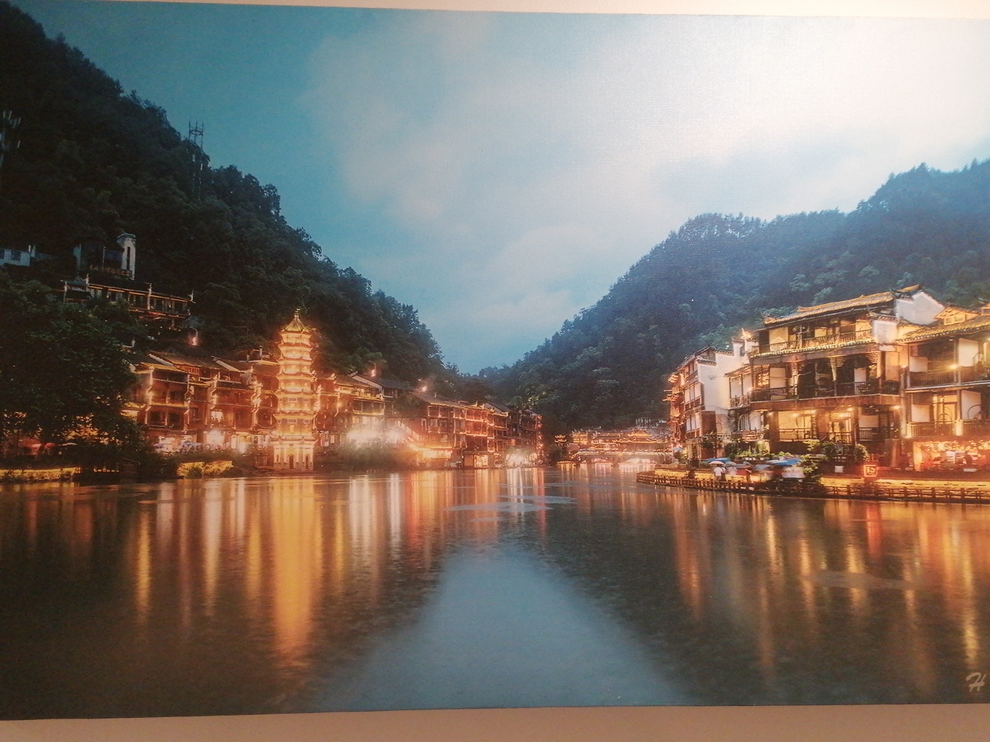 The image depicts a cityscape at dusk, with buildings lining a river that reflects the warm light of the structures. Buildings are mostly multi-story structures with traditional architecture, appearing to be lit from within. A pagoda-like tower stands prominently among the buildings on the left bank. Dense, green hills frame the scene on both sides of the river. People are not clearly visible, but there are signs of activity along the riverbank. The light suggests early evening. The location is likely not Sa Pa Ward, Vietnam, as the architecture is Chinese, not Vietnamese.