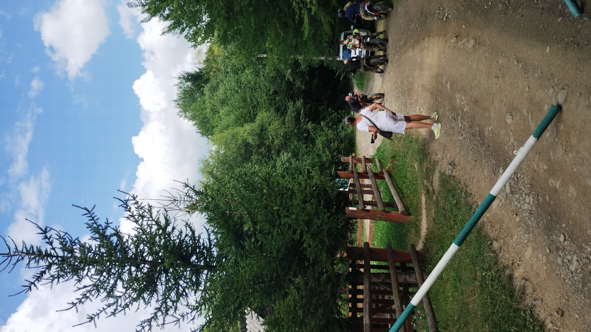 In an outdoor, rural setting, several people are gathered around ATVs and a white van on a dirt road. The road is lined with a wooden fence and lush green trees. A large pine tree stands prominently in the foreground. A person in a white shirt and shorts is facing away from the camera, holding a phone. Another person is on an ATV, wearing a helmet. The sky above is blue with scattered white clouds. A white and green striped barrier is laid across the dirt road in the foreground.