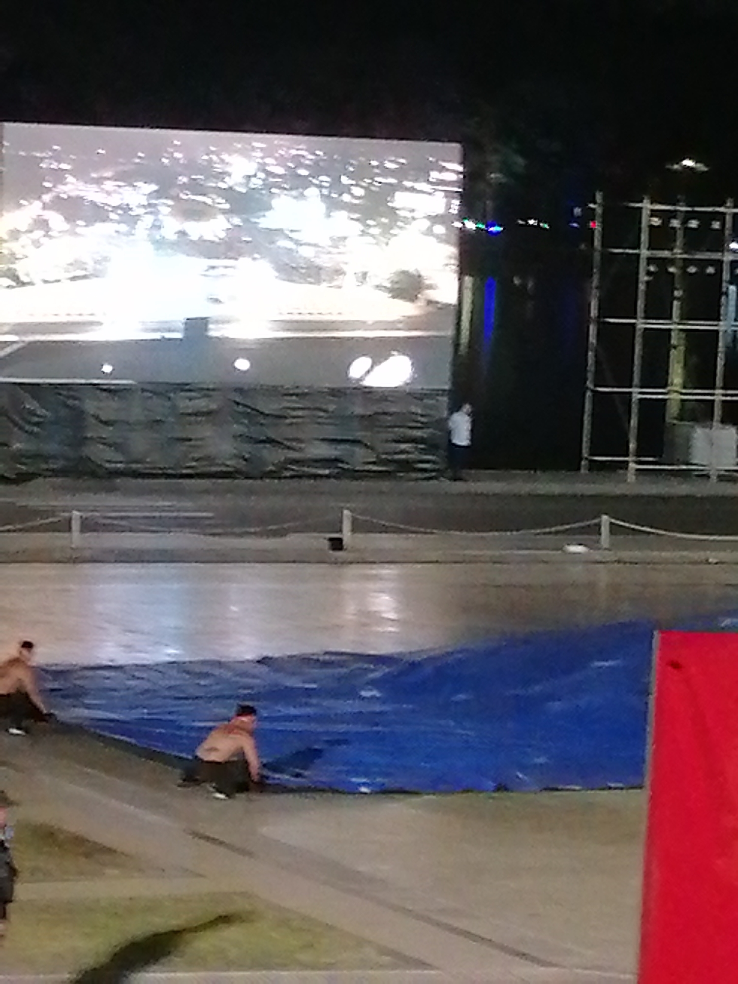 In Da Lat, Vietnam, the image depicts an outdoor scene at night with a large screen displaying bright lights. In the foreground, two shirtless men are kneeling on a blue tarp laid out on a sloped surface. One man is in the process of pulling on the tarp, while the other appears to be adjusting it. To the right, a portion of a red object is visible. In the background, a metal scaffolding structure stands next to the screen. A person in a white shirt is standing near the screen. The environment appears to be an open area, possibly a performance venue or event space.