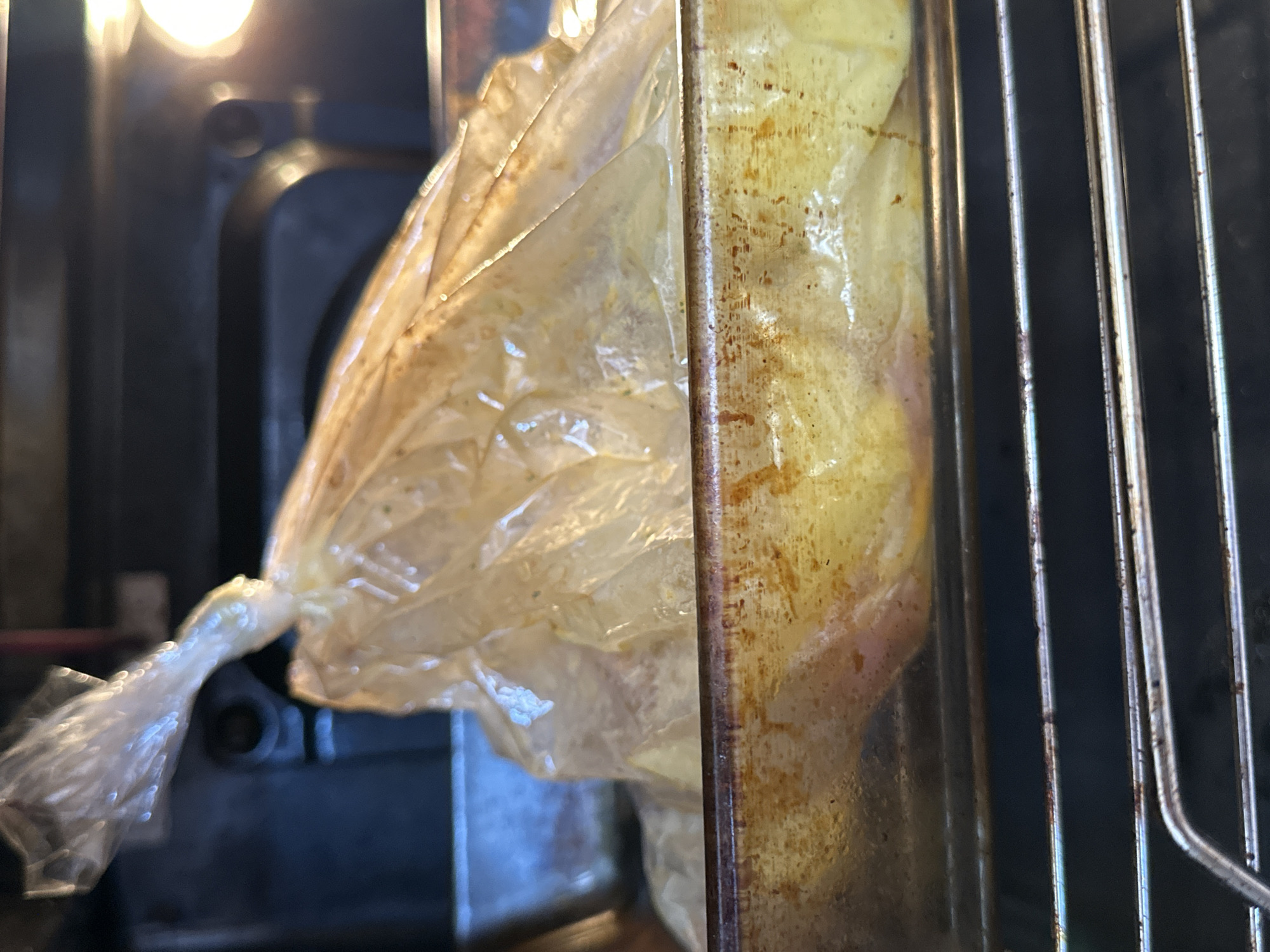 A close-up shot inside an oven shows a clear plastic bag containing an item with a yellow hue. The bag is tied at the top, obscuring the contents. The oven rack is visible in the right portion of the frame and appears tarnished. The background reveals part of the oven's interior, which is dark and indistinct. The location is not discernible from the image.