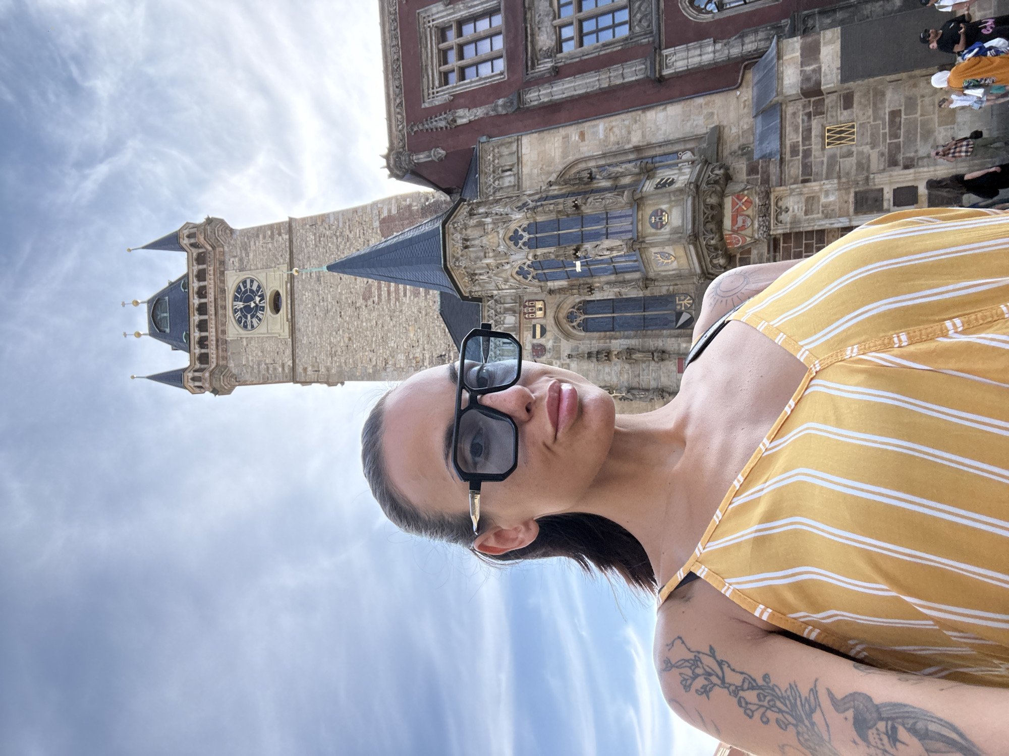 A woman with dark hair pulled back and wearing large sunglasses is taking a selfie. She has tattoos on her left arm and shoulder and is wearing a yellow and white striped dress. Behind her, the Old Town Hall Tower in Prague, Czechia, is visible. The tower has a large clock face and spires. Part of a red building with ornate windows is also visible to the right of the tower. Several people are walking in the background, and the sky is cloudy.