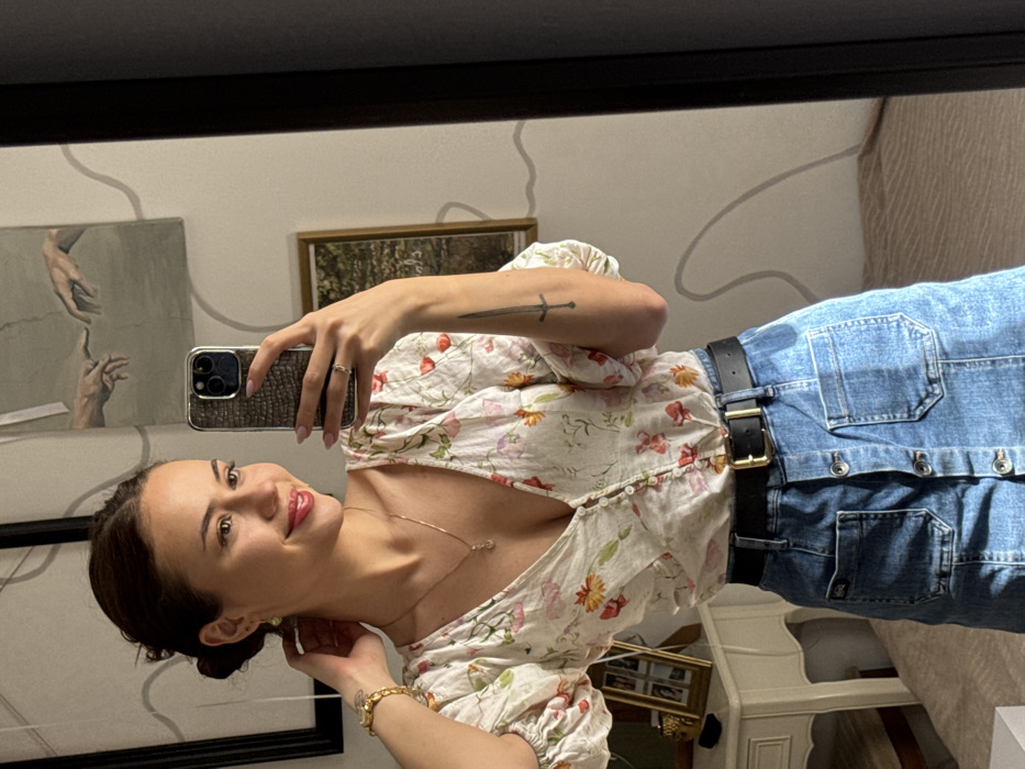 An indoor mirror selfie captures a young adult female, approximately 20-35 years old, within a domestic interior in Tel Aviv-Yafo, Israel. The female subject has dark hair styled in an updo, a light complexion, and red lip makeup. She wears light green stud earrings, a thin gold necklace with a small pendant, and a gold watch on her left wrist. A sword tattoo is visible on her inner left forearm. She is dressed in a light-colored, V-neck blouse with a floral pattern featuring red, orange, and green elements, paired with a medium-wash denim skirt with a button-front closure and two large patch pockets. A black belt with a gold rectangular buckle secures her waist. Her right hand, featuring light pink fingernail polish and a gold ring on the ring finger, holds an iPhone 15 Pro/Pro Max in a dark, textured phone case, pointed towards the mirror to capture the image. Her left hand is positioned near her left ear. The background, visible through the mirror's reflection, consists of a light-colored wall with wavy gray lines, a large black-framed print depicting the hands from Michelangelo's "The Creation of Adam," a smaller framed landscape print, and a light-colored vanity table holding a gold multi-photo frame and a gold decorative object. A light-colored patterned rug is partially visible on the floor. Natural light illuminates the scene from the left.